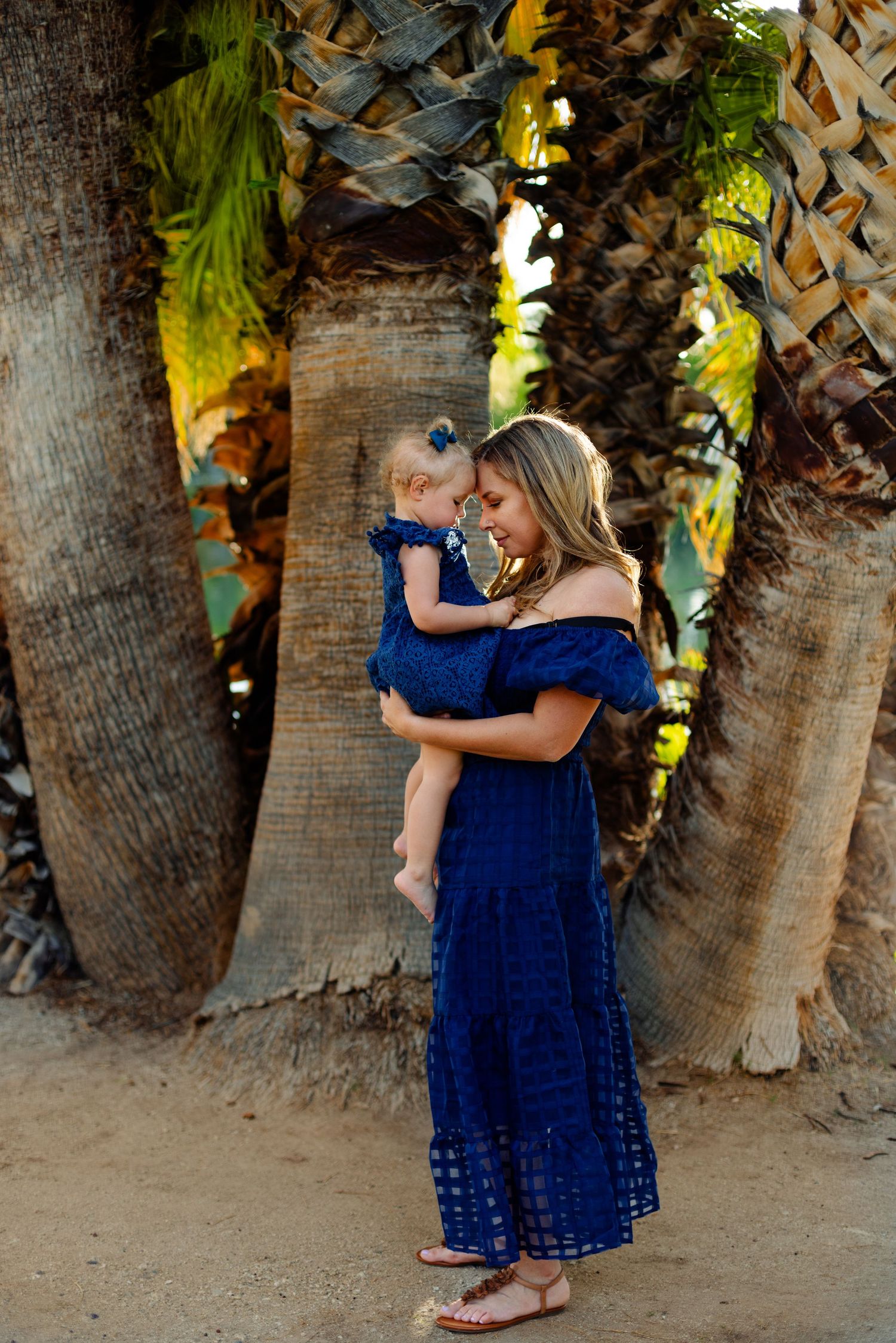 Two children in blue dresses embrace near palm trees at sunset.
