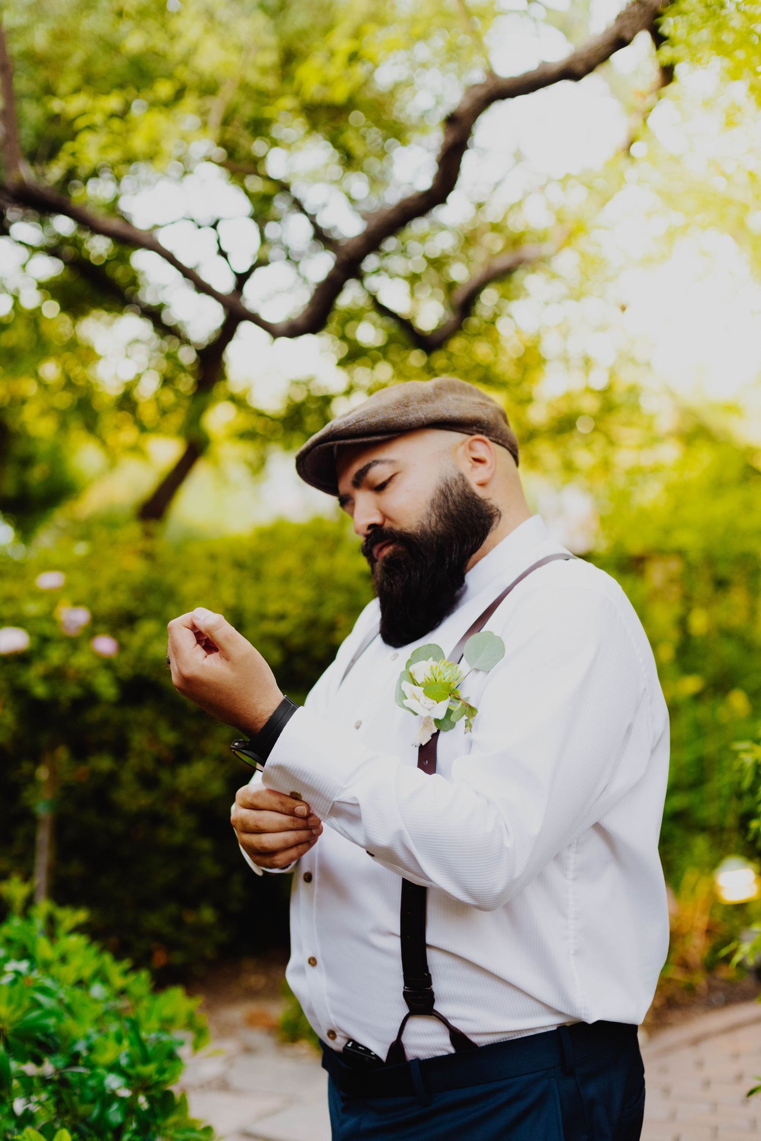 A formal portrait of a groom wearing a flat cap and white dress shirt with suspenders in a garden setting.