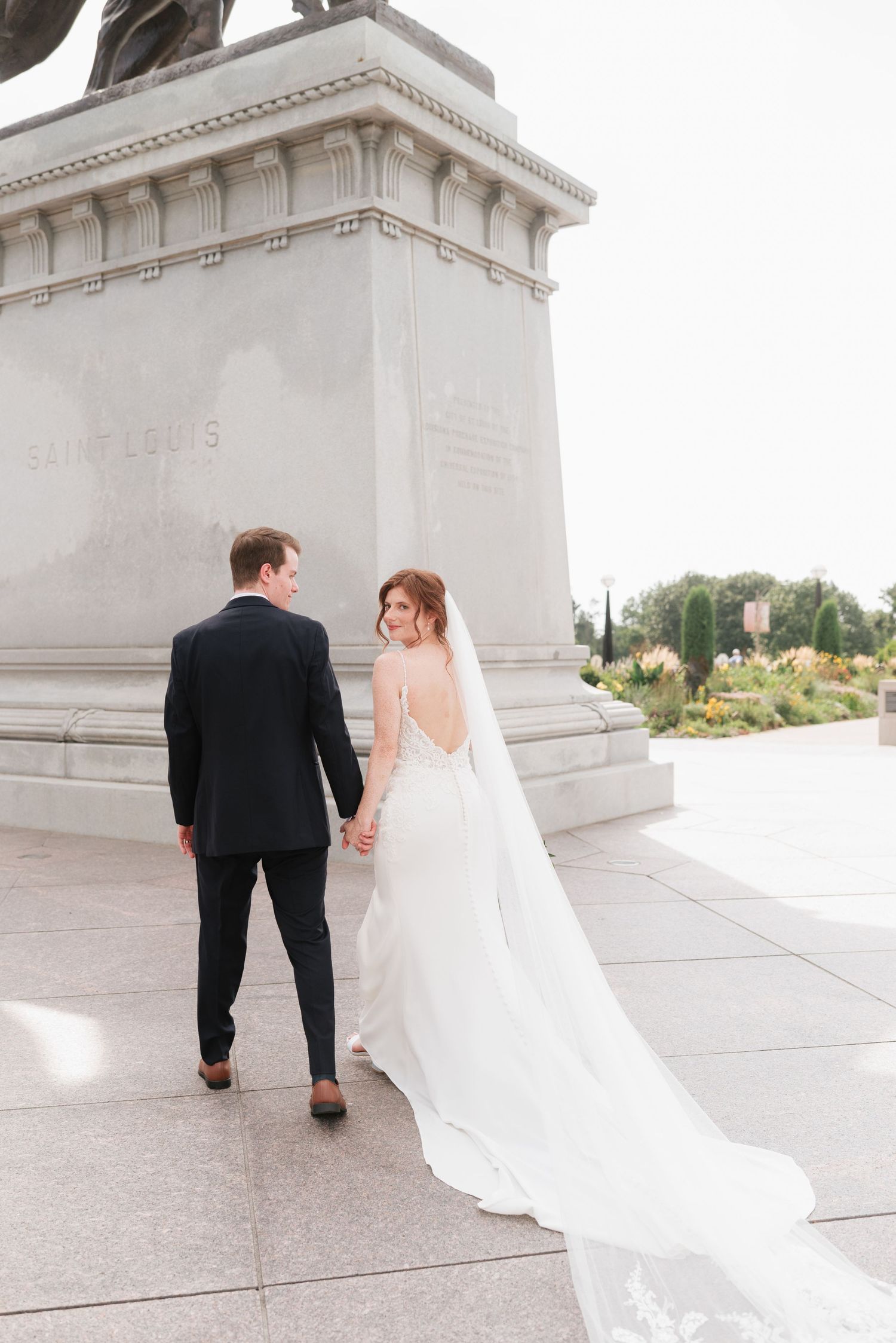 Bride and groom walk hand-in-hand near a marble monument, her flowing veil trailing behind on a sunny wedding day.