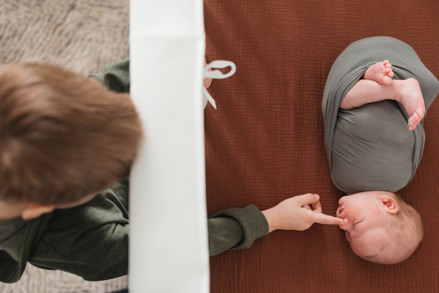 Older sibling gently touching newborn baby wrapped in gray swaddle on rust brown blanket, aerial view.