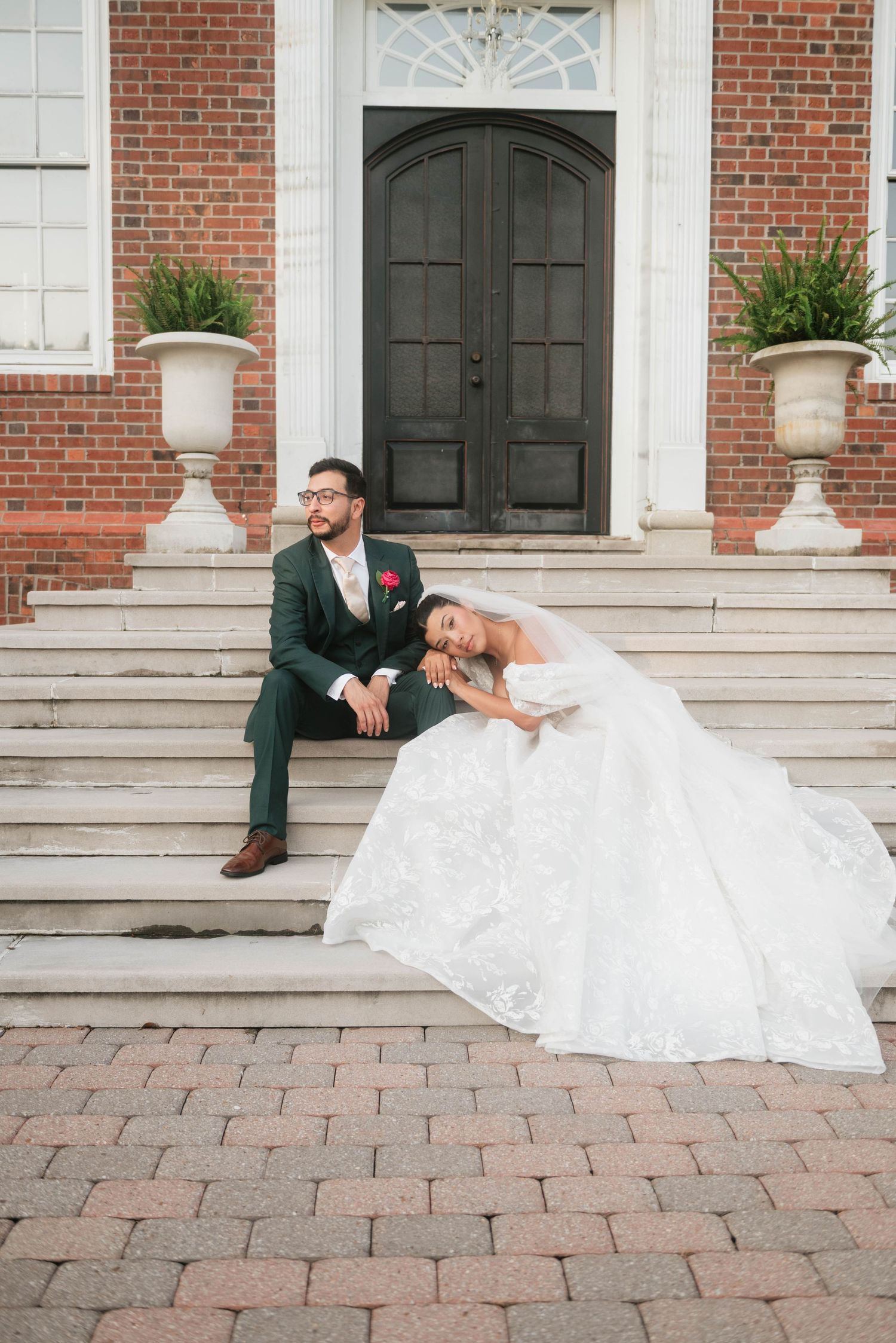 Bride in white gown leans on groom in green suit on brick steps of elegant building on wedding day.