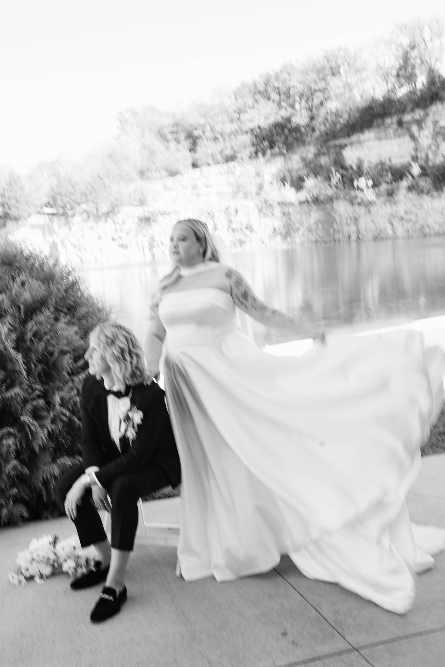 Black and white wedding photo of bride in flowing gown and groom in tuxedo posing by a serene waterfront.