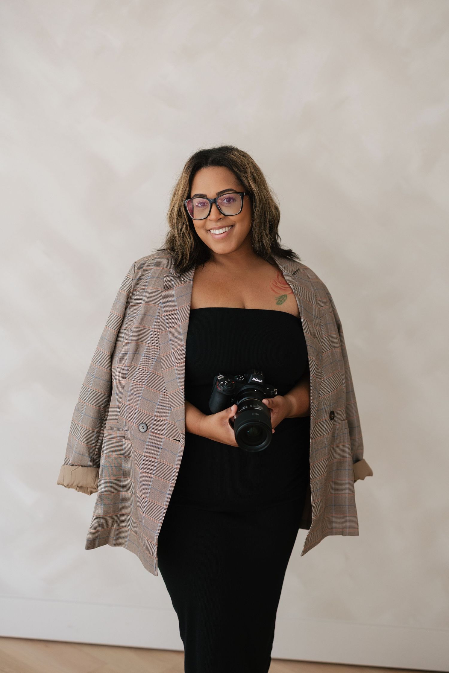 Smiling woman in black dress, plaid blazer, and glasses holds a camera, posing confidently against a neutral background.