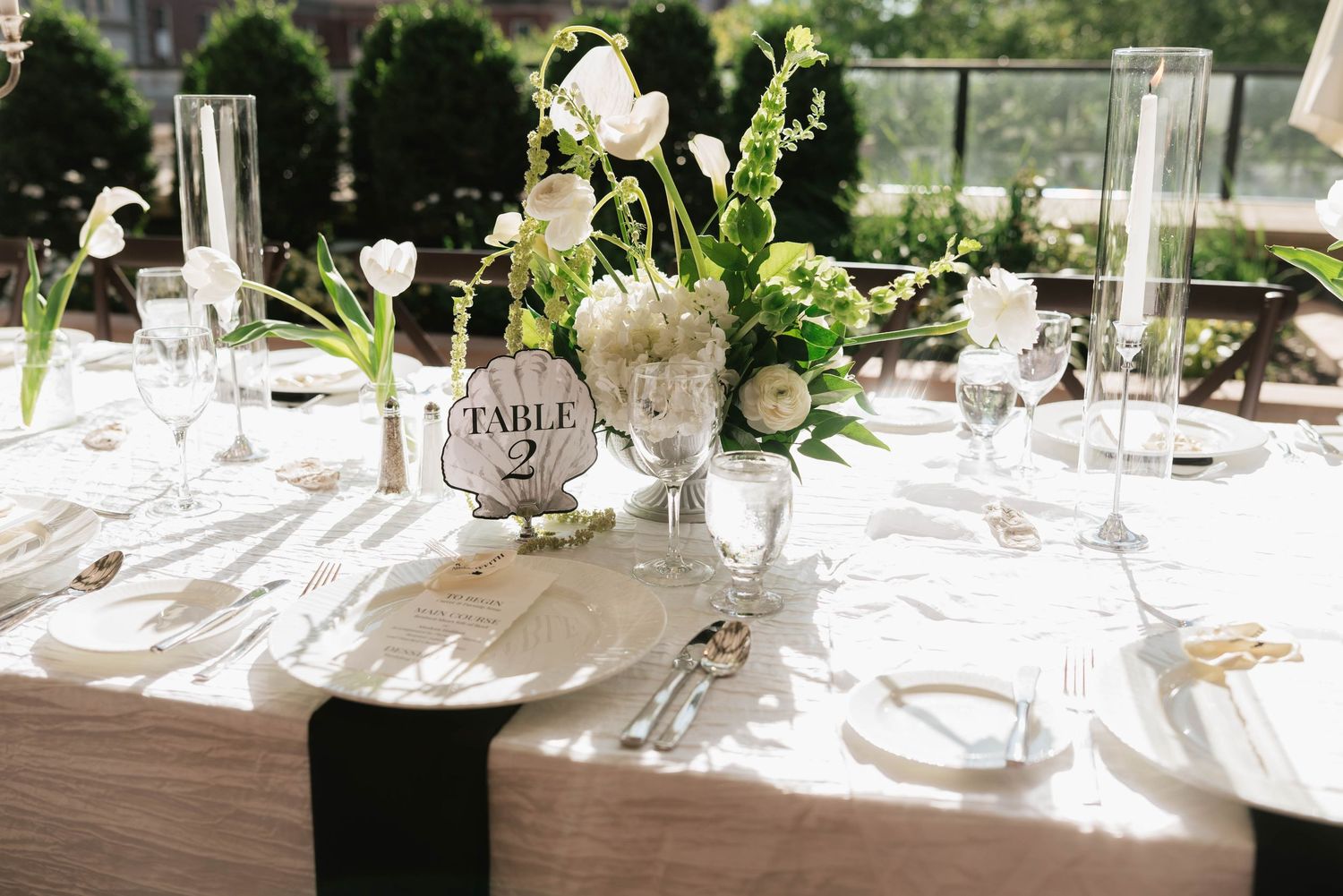 Elegant wedding reception table setting with white floral centerpiece, Table 2 sign, crystal glassware, and black napkins.