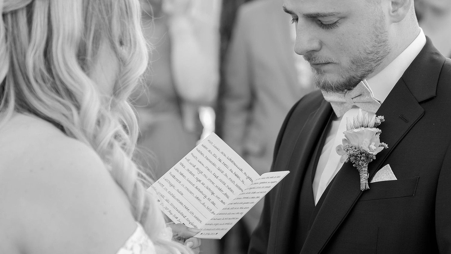 A groom in a black suit reads handwritten vows during an emotional wedding ceremony moment.