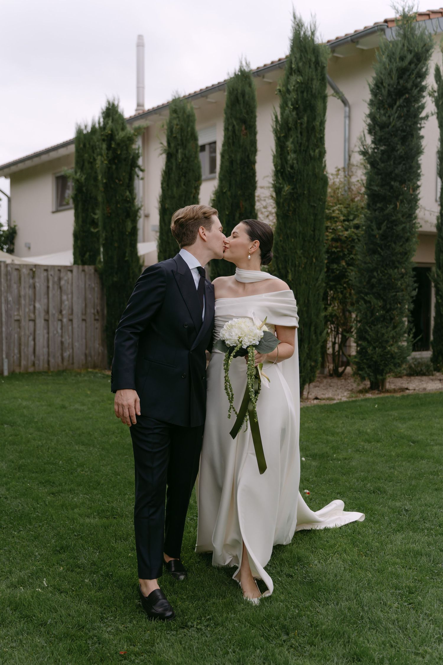 Wedding couple posing in garden with tall cypress trees and white house backdrop.