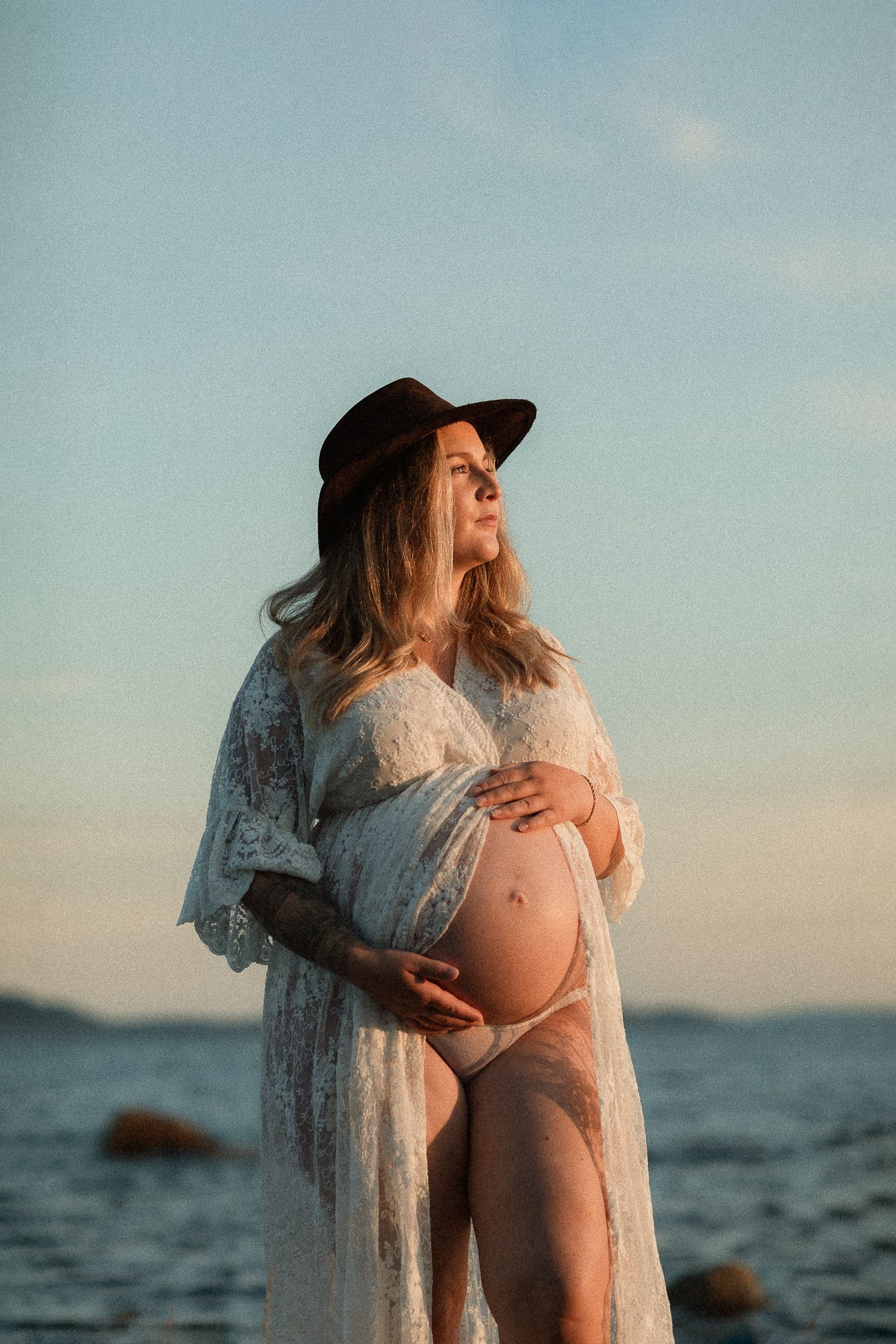 En gravid kvinna i spetsklänning och cowboy hatt, vid havet och med en blå himmel i bakgrunden.