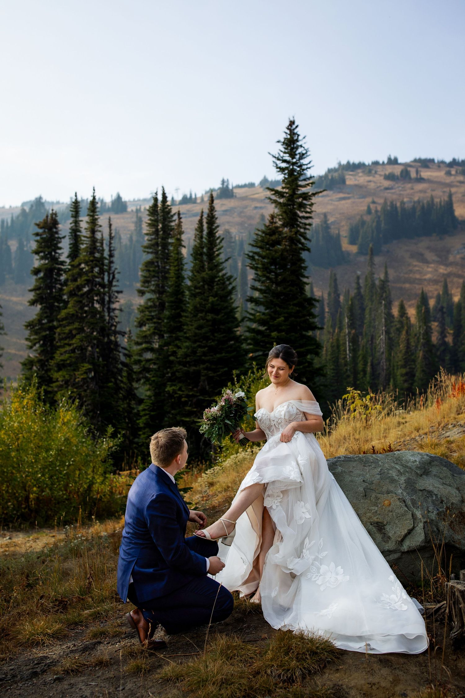 Groom fixes brides shoe during their wedding portraits at Sun Peaks Resort among the mountains and summer season colours.