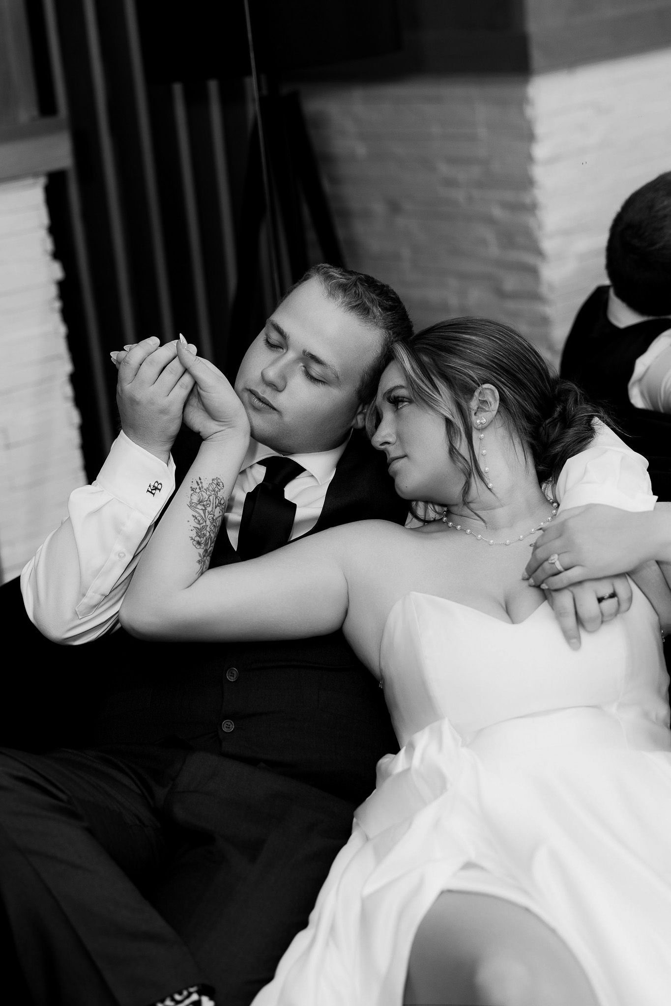 Black and white moody wedding portrait of bride and groom cuddling on a couch, groom gently leaning to kiss bride’s hand, capturing intimate