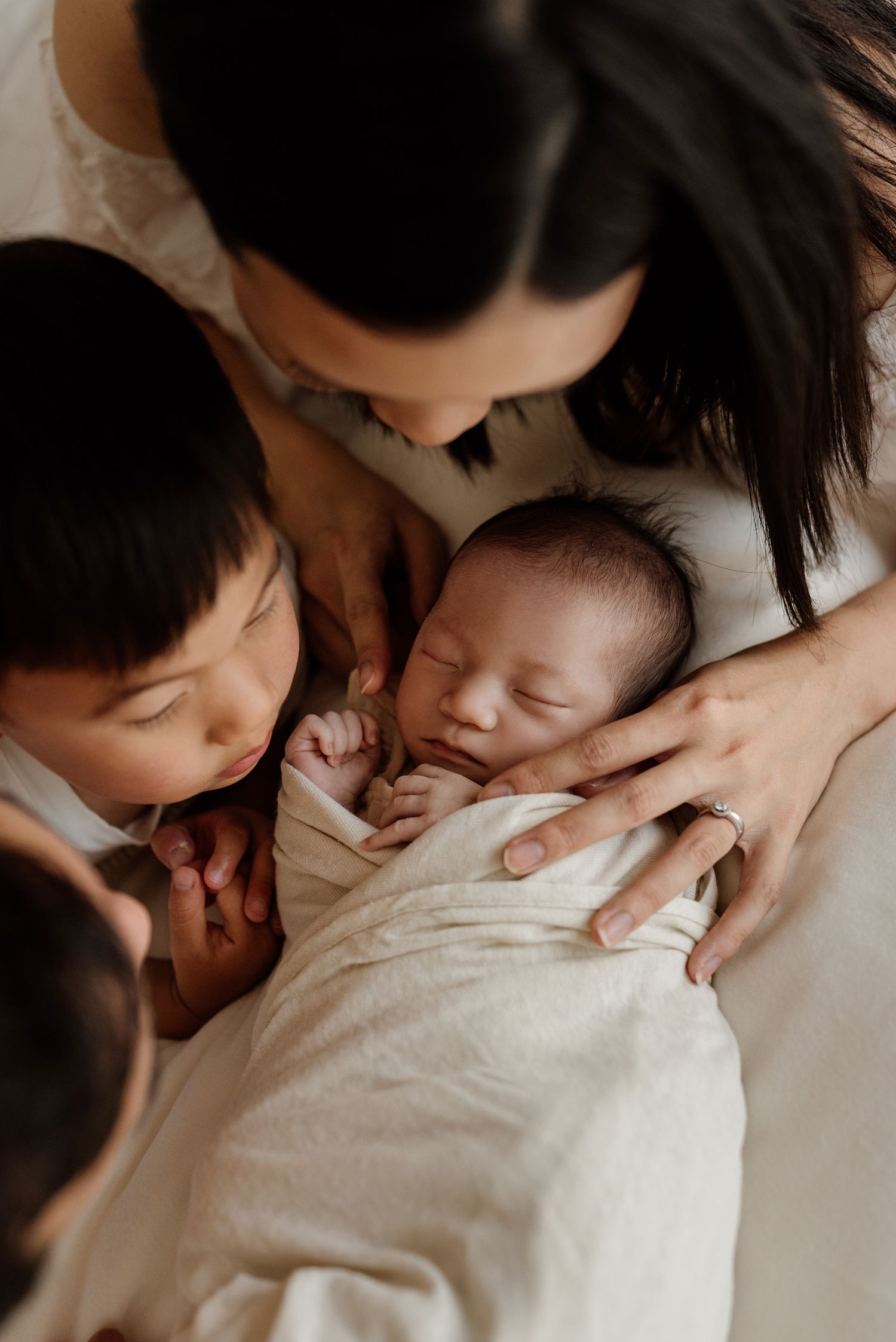 Mother and sibling lovingly gazing at sleeping swaddled newborn baby during family photo session.