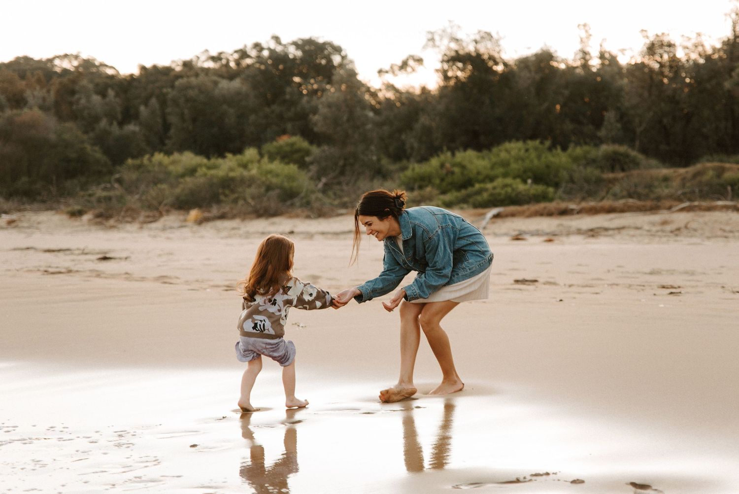 A mother and young daughter playing together on a sandy beach at sunset, sharing a joyful moment by the water in Sydney.