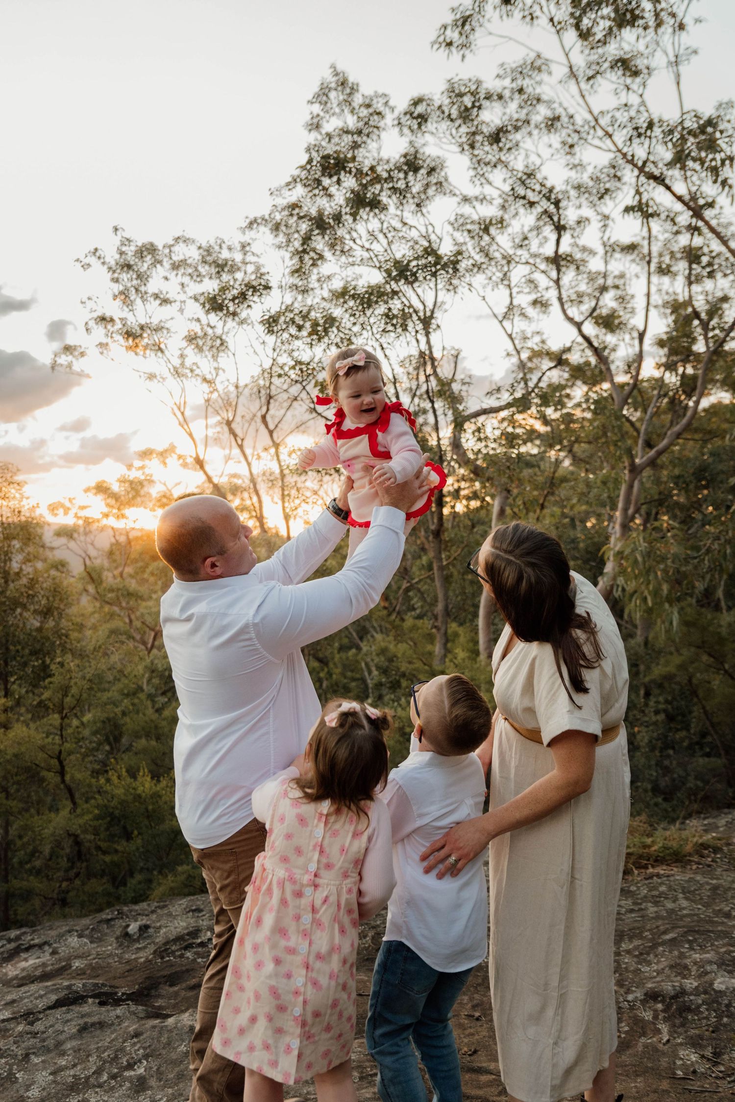 Family of five enjoying a golden sunset outdoors, father lifting baby girl while children gather around mother on rocky terrain.