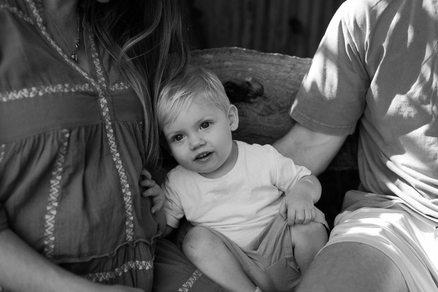 Black and white photo of a smiling baby sitting between two adults outdoors.