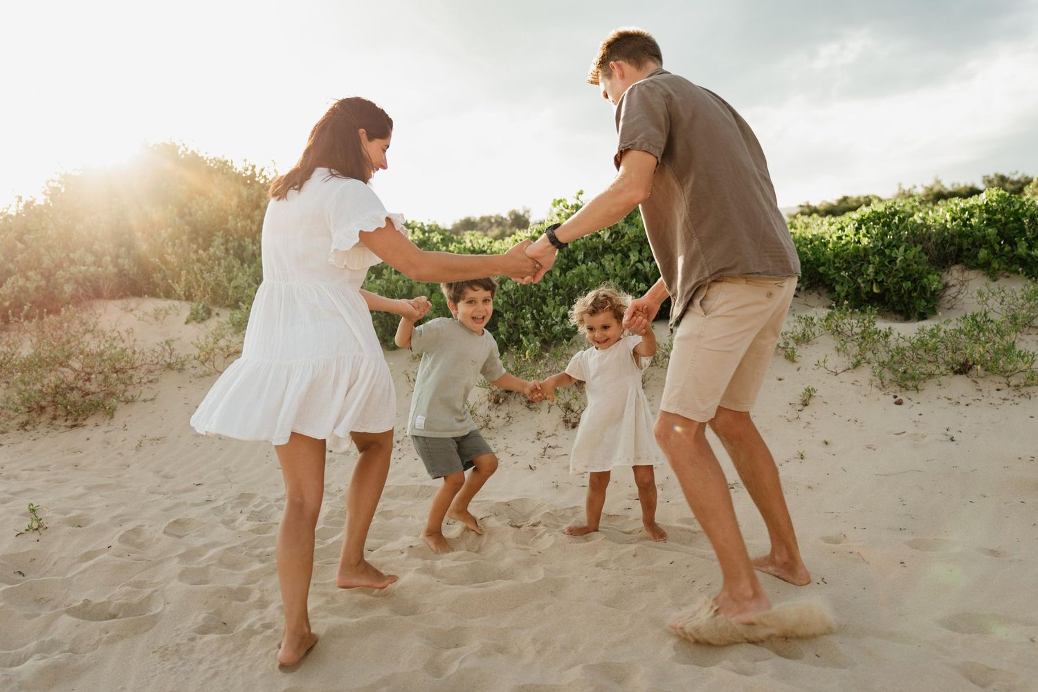 A joyful family of four holding hands and playing in a circle on a sandy beach at sunset.