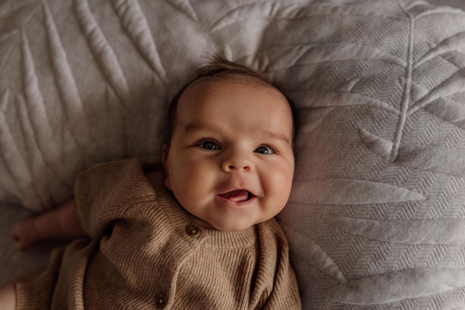 Smiling newborn baby in a brown knit romper lying on a soft gray textured blanket.
