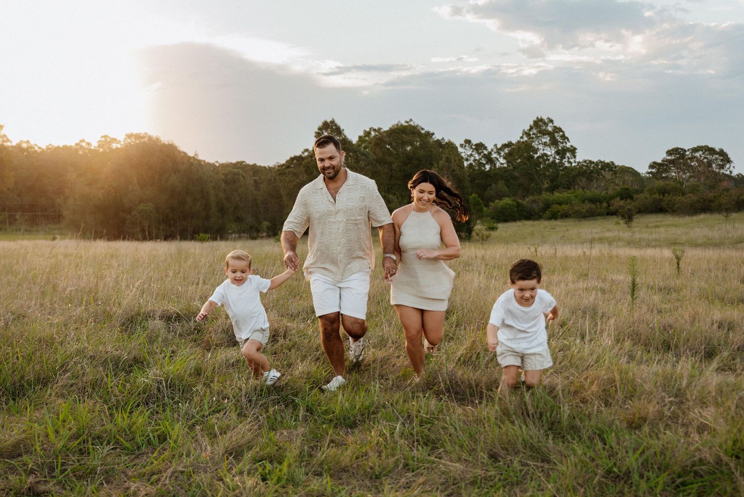 Family of four in white outfits running through a golden sunlit field at sunset.