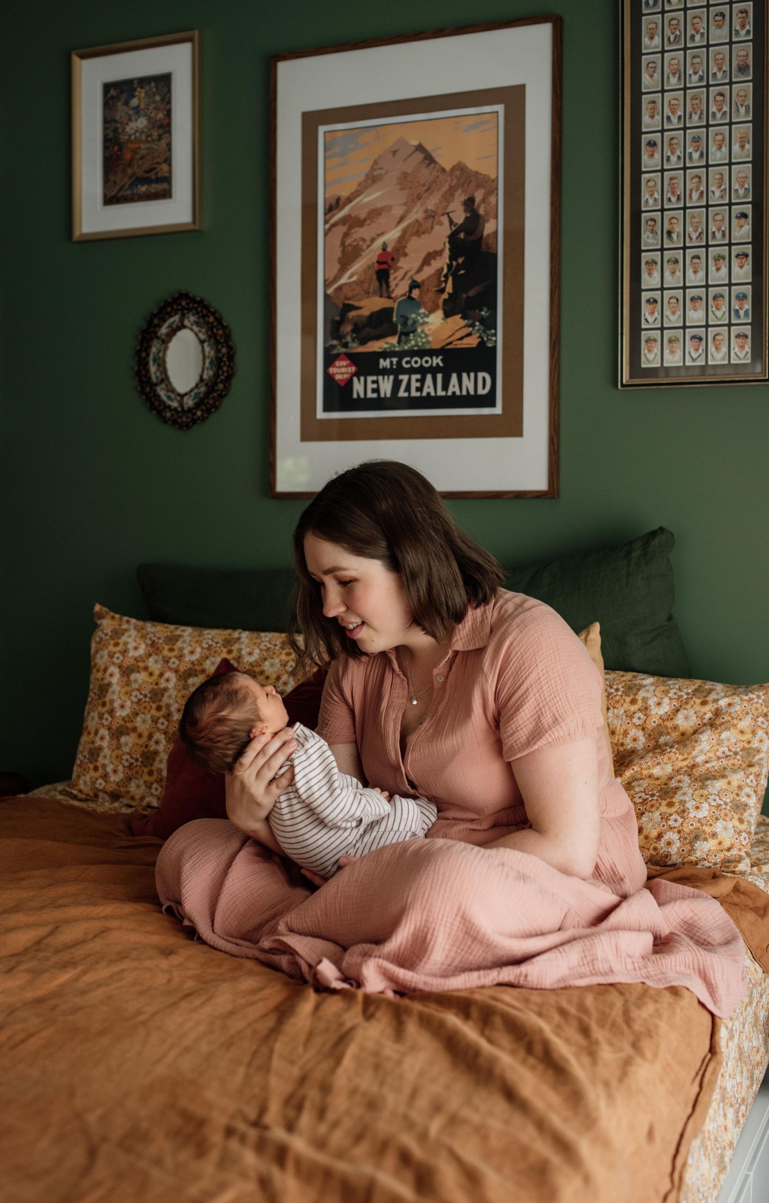 Mother in pink dress cradles newborn on bed with mustard linens and New Zealand poster backdrop.