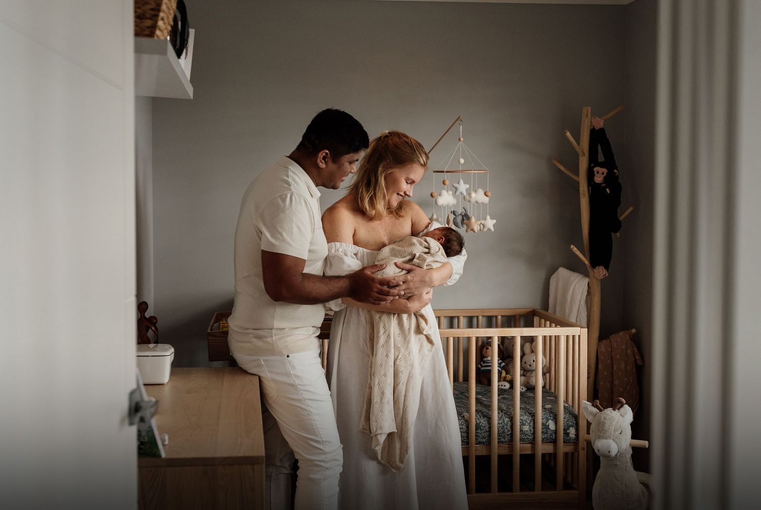 Couple in white outfits admiring their newborn baby in a cozy nursery with a wooden crib.