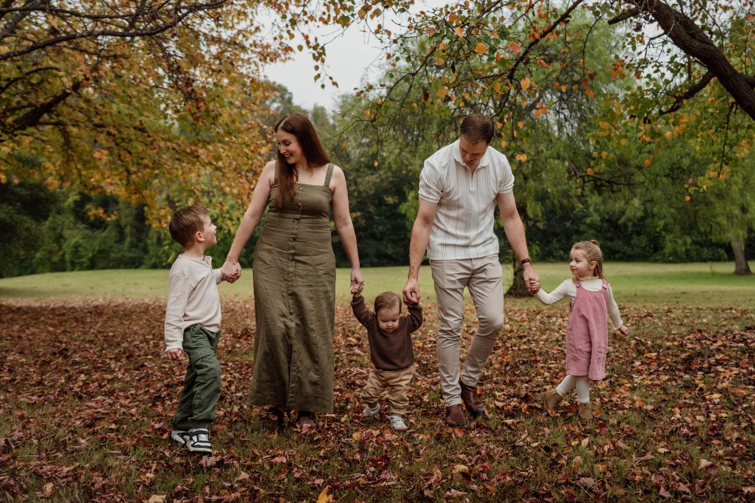 Family of five walking through autumn leaves at Tench Reserve Nepean River, parents holding toddler's hands while two children smile.