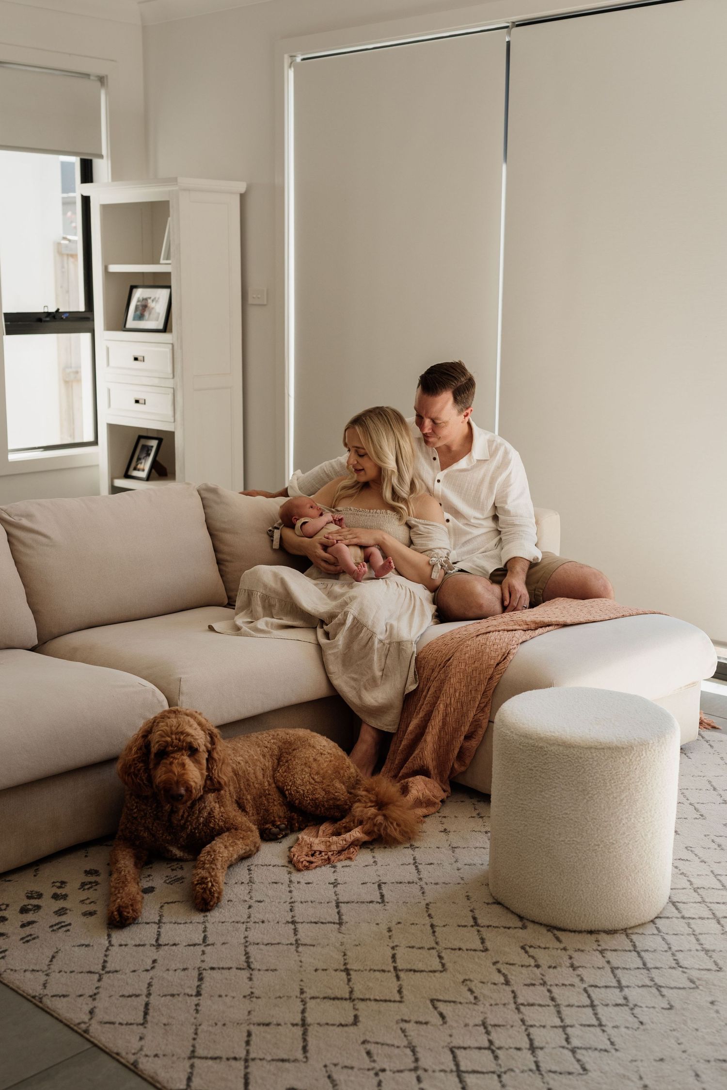 Couple sitting on beige sofa with newborn baby, golden doodle dog lying on patterned rug in bright living room.