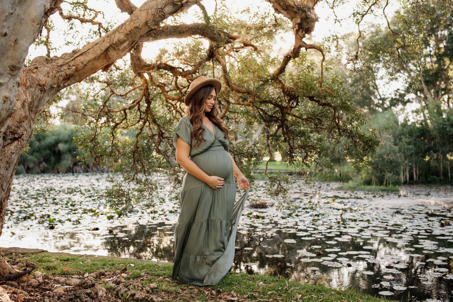 Pregnant woman in green maxi dress poses under tree by lily pad pond for maternity photo session.