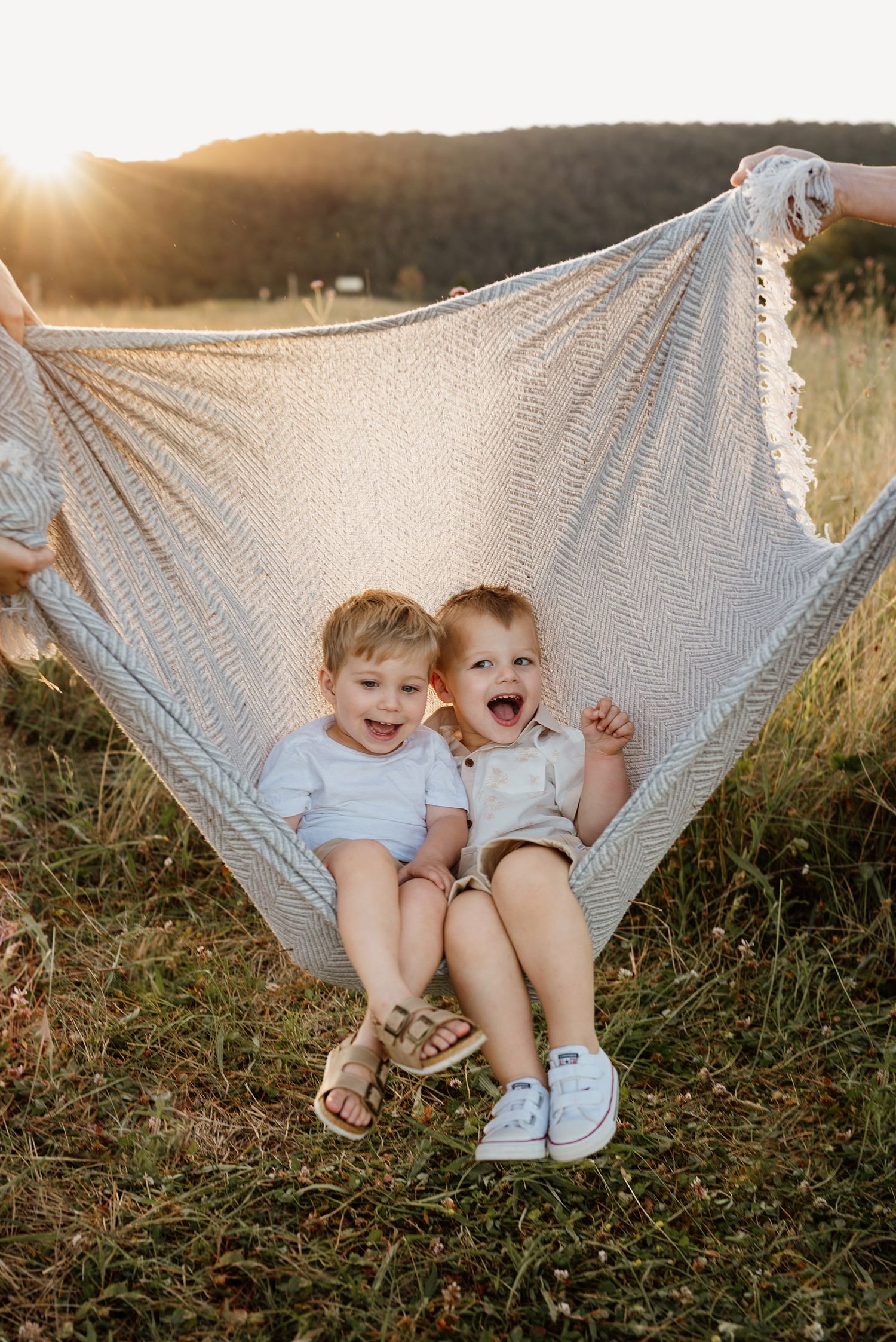 Two happy toddlers laughing in a hammock outdoors at golden hour, wearing white outfits in a sunny meadow.