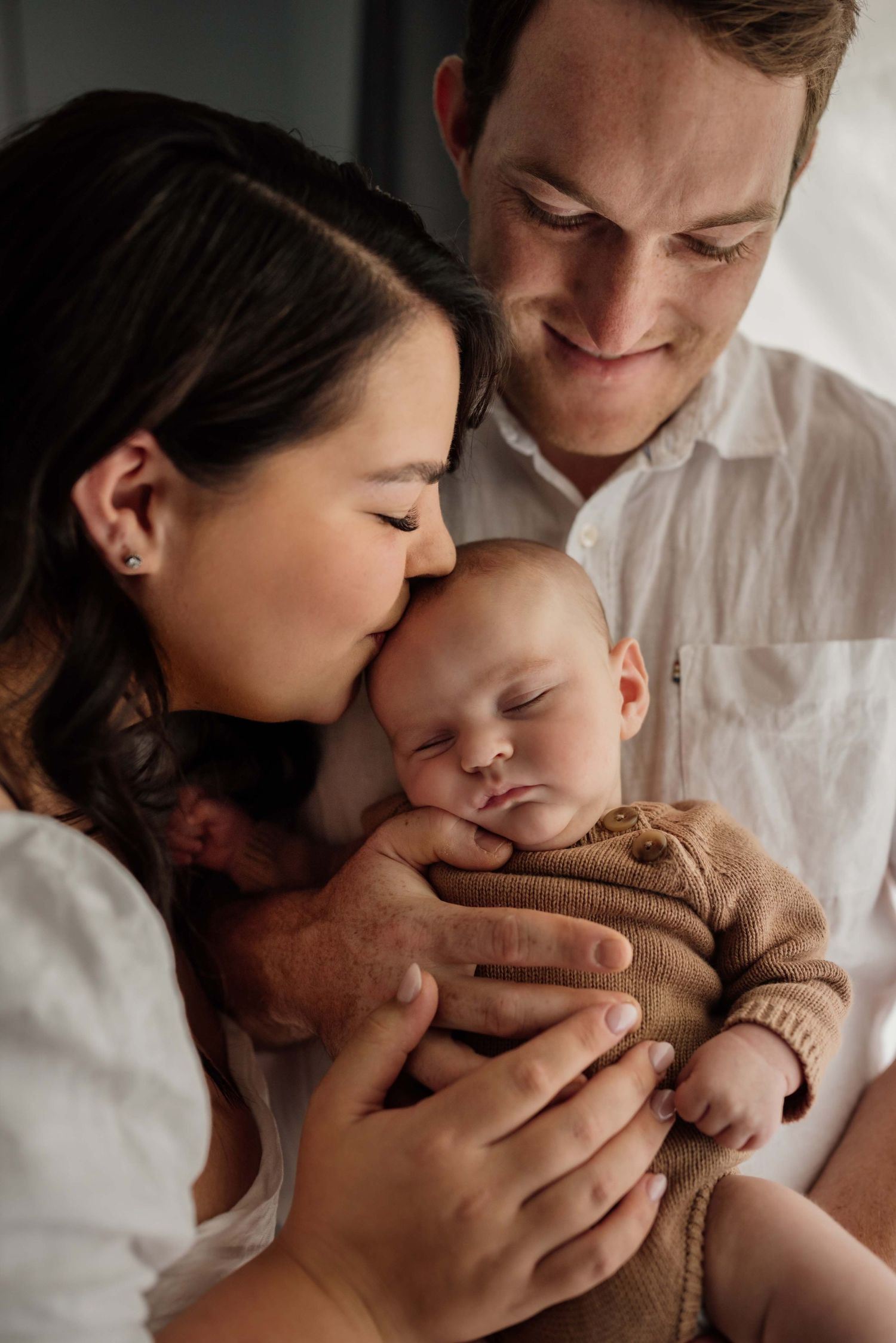 Loving parents in white clothing gently kiss and hold their sleeping newborn baby.