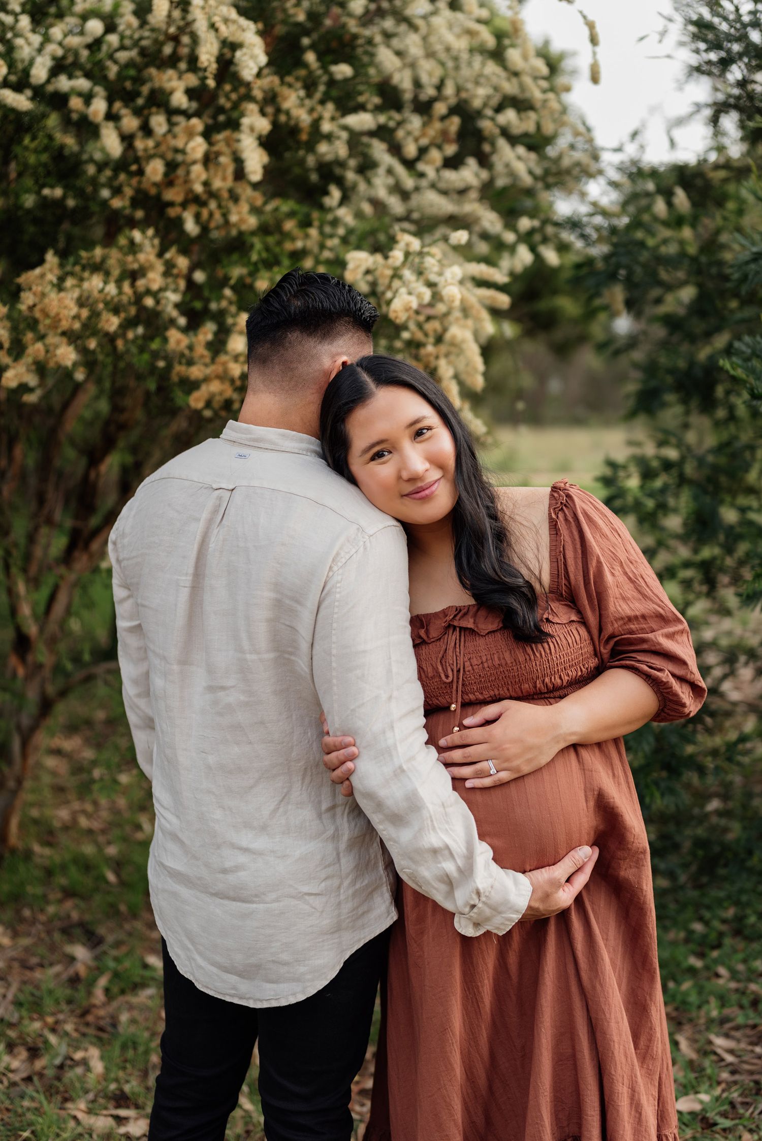 Couple sharing a tender maternity portrait outdoors, woman in rust dress cradling baby bump amid blooming white flowers in Penrith.