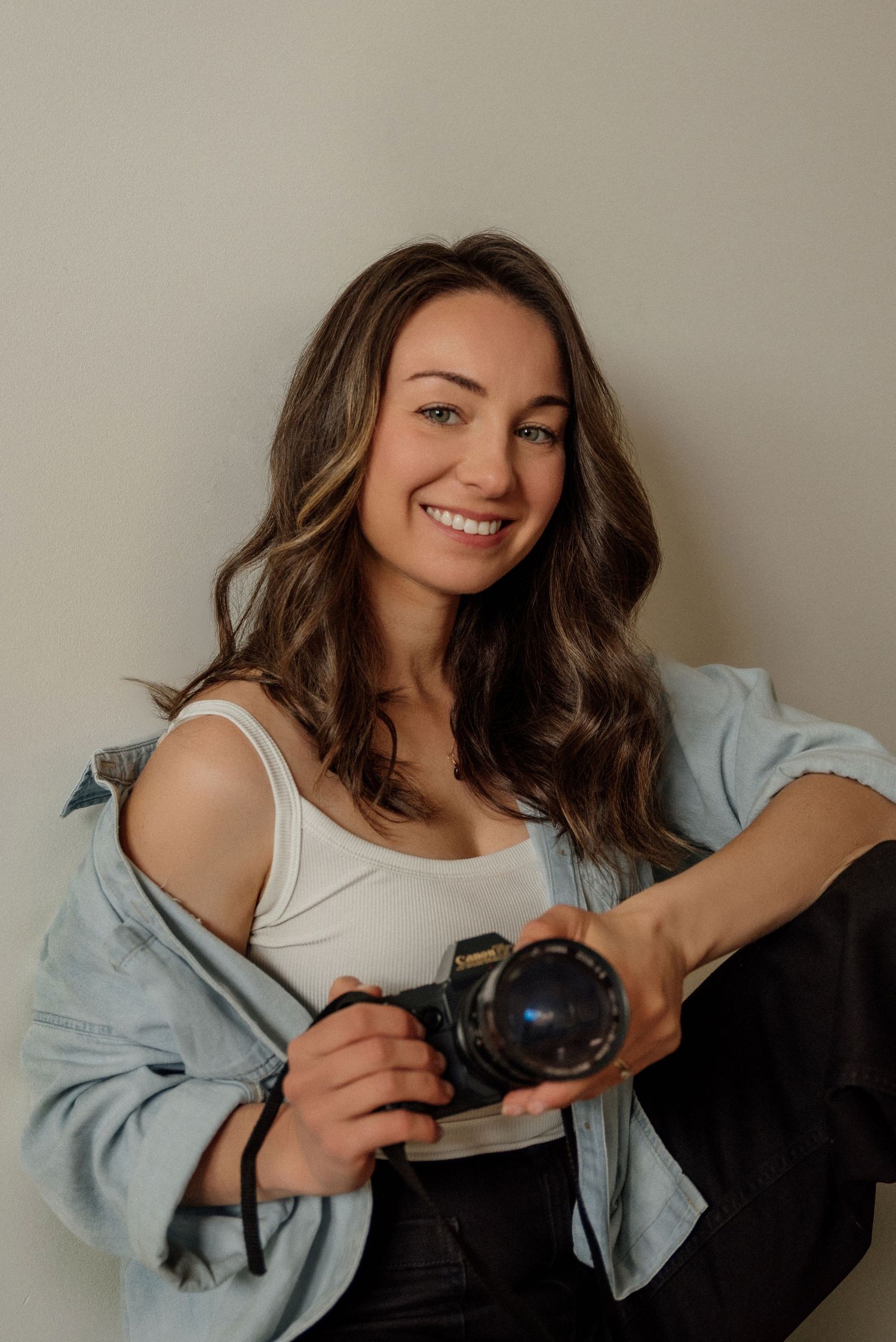 Smiling young female photographer holding a camera while sitting against a neutral wall.