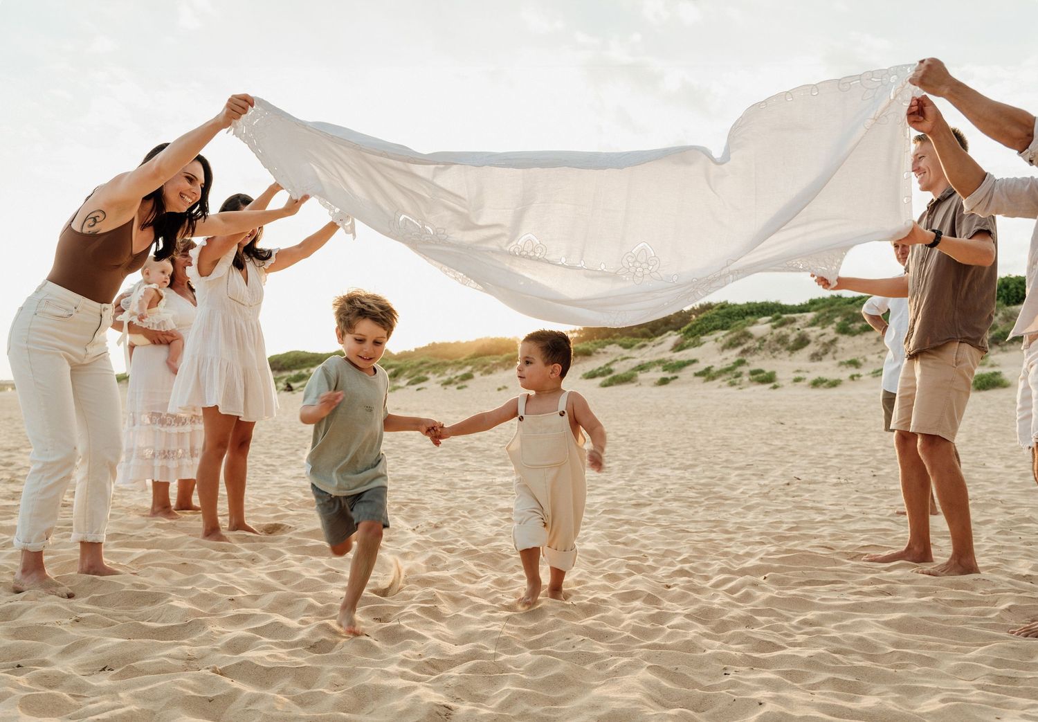 Extended family plays with white sheet over two young children running on sandy beach.