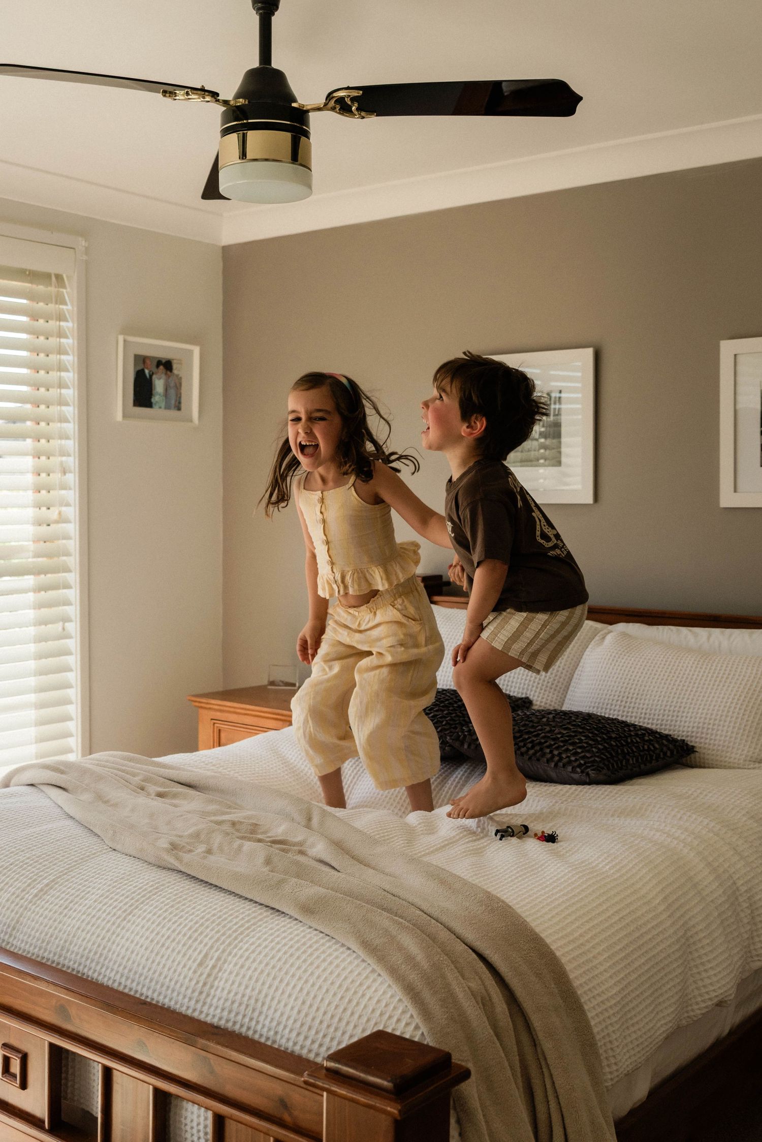 Two children joyfully jumping on a bed in a cozy bedroom with neutral decor and a ceiling fan in Penrith.
