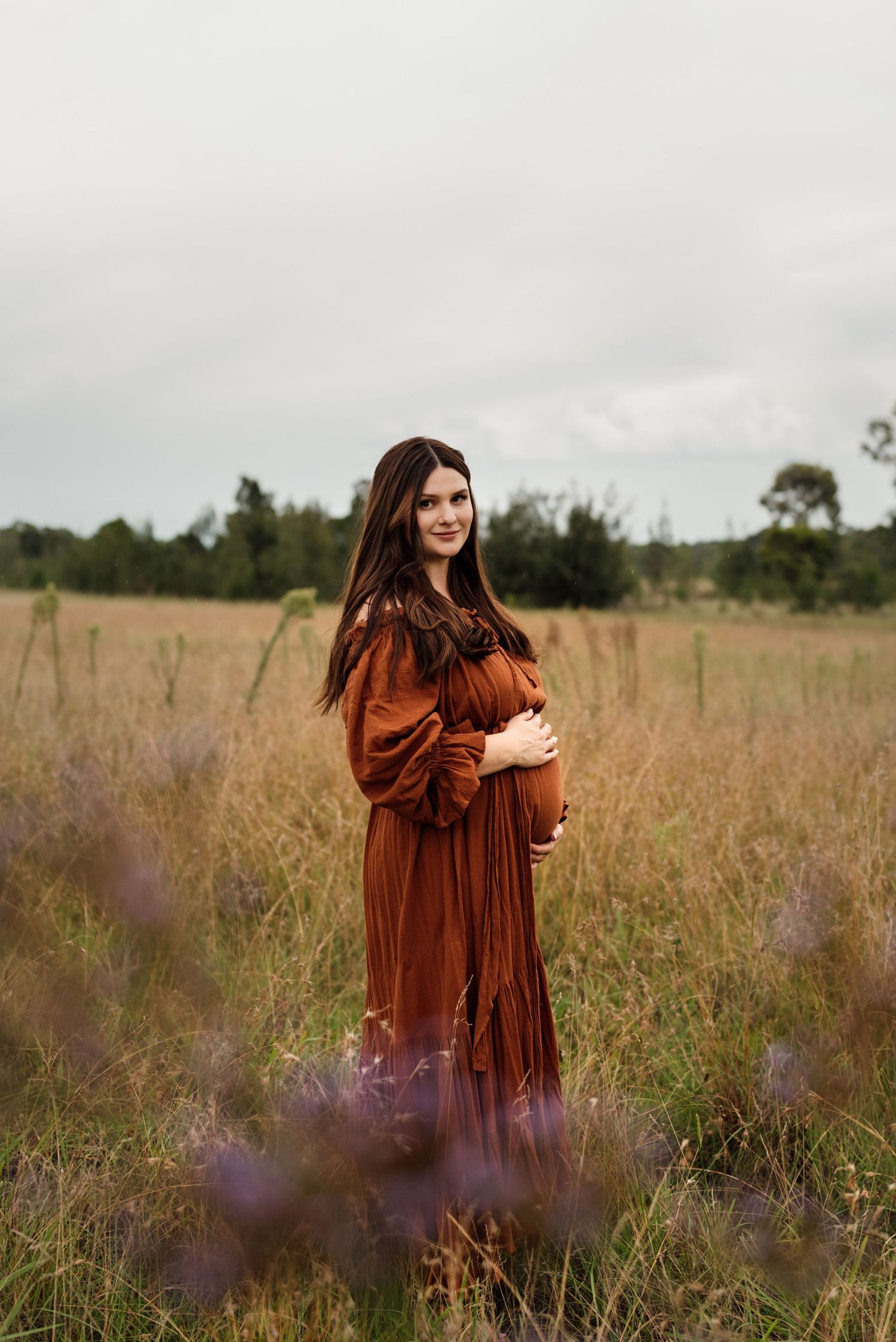 Pregnant woman in a flowing rust-colored maternity dress cradles her belly while standing in a golden grass field under an overcast sky.