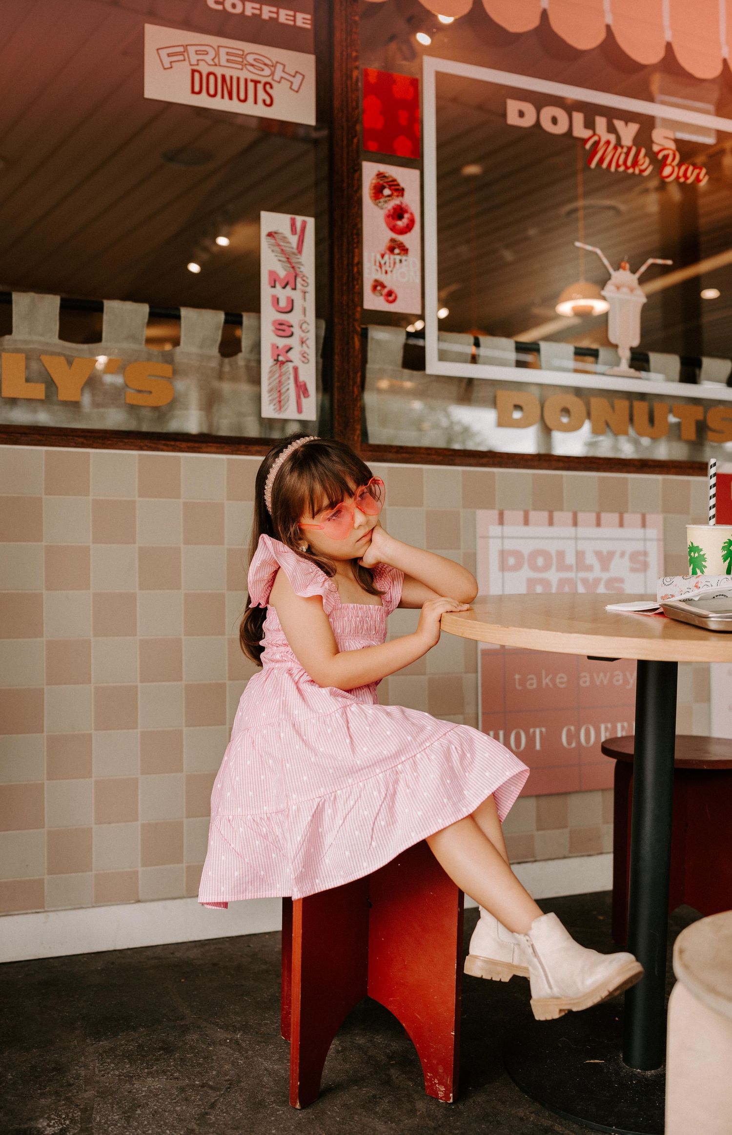 Young girl in pink dress and heart sunglasses poses at a retro diner table inside Dolly's Milk Bar donut shop.