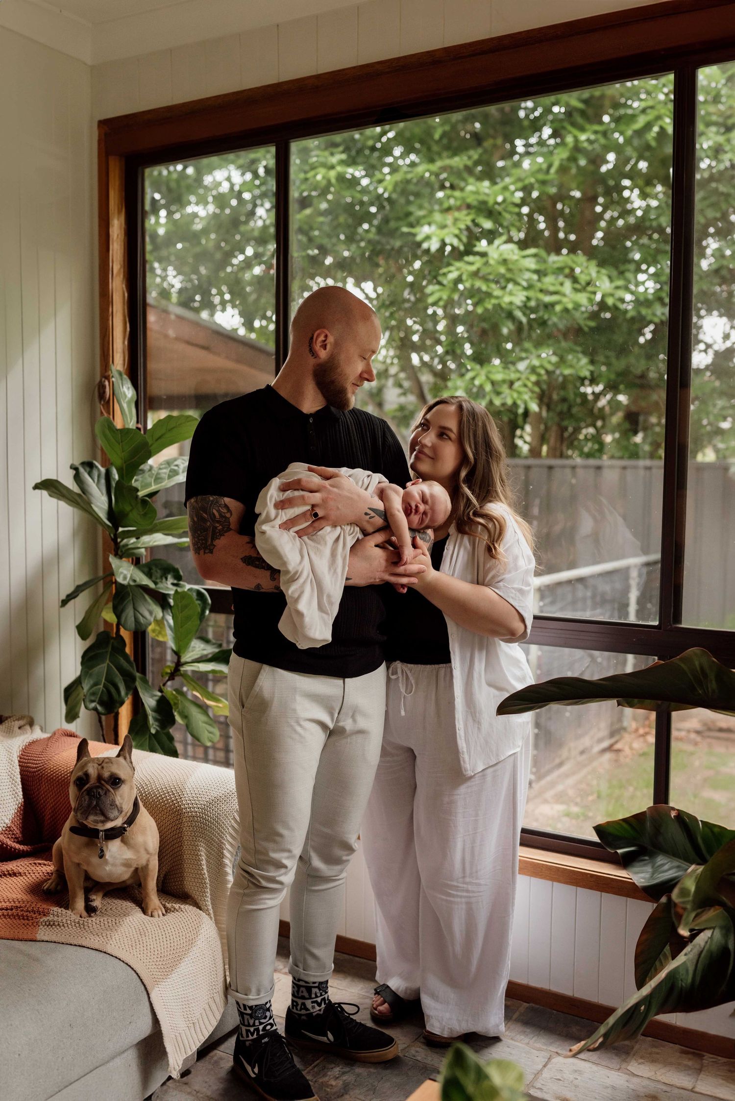 Young couple holding newborn baby in sunlit living room with French bulldog and tropical plants.