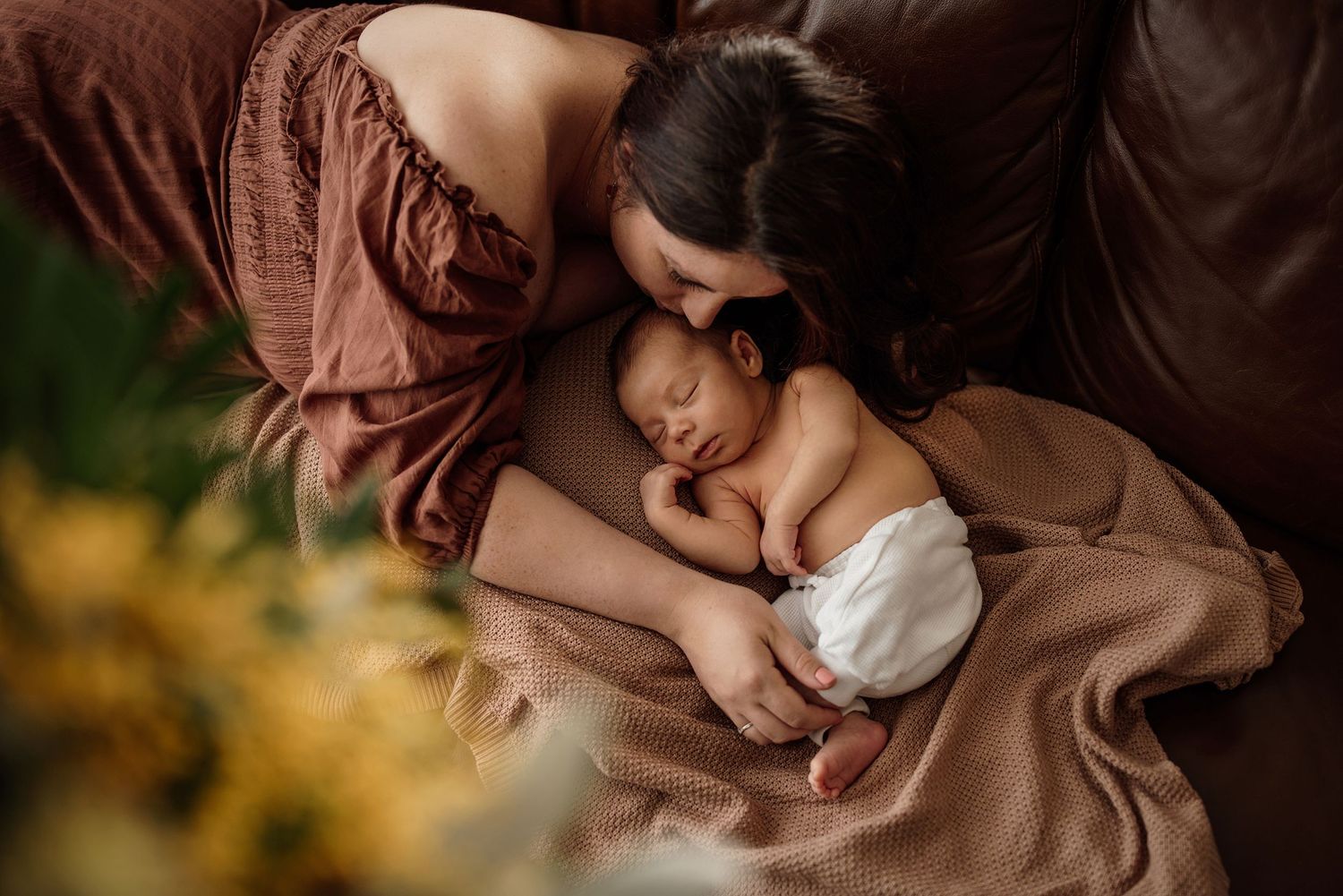 A mother tenderly kisses her sleeping newborn baby wrapped in a soft brown blanket during a newborn photography session.