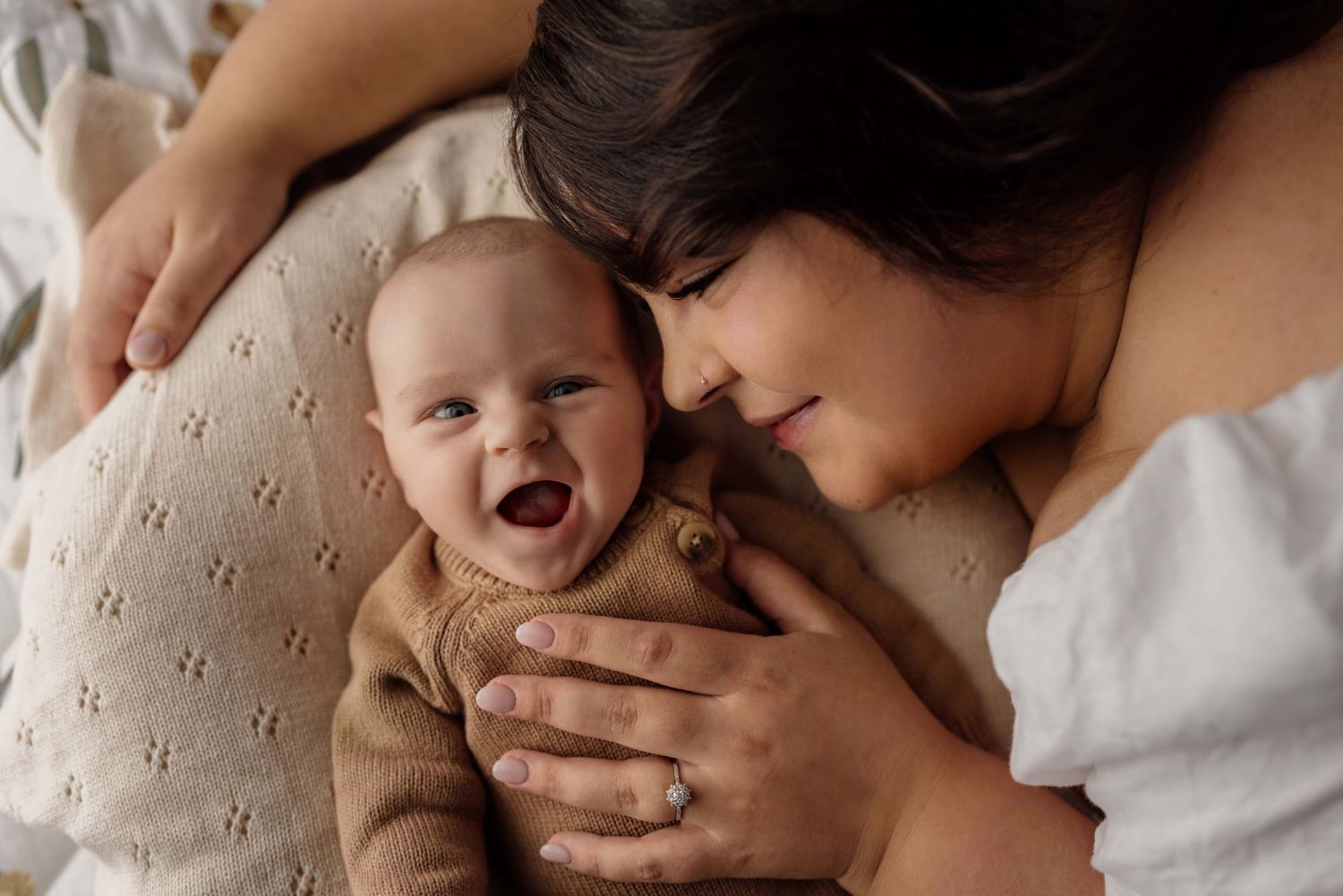 A smiling mother lying down gazing at her laughing newborn baby dressed in a brown knit onesie on a soft bed.