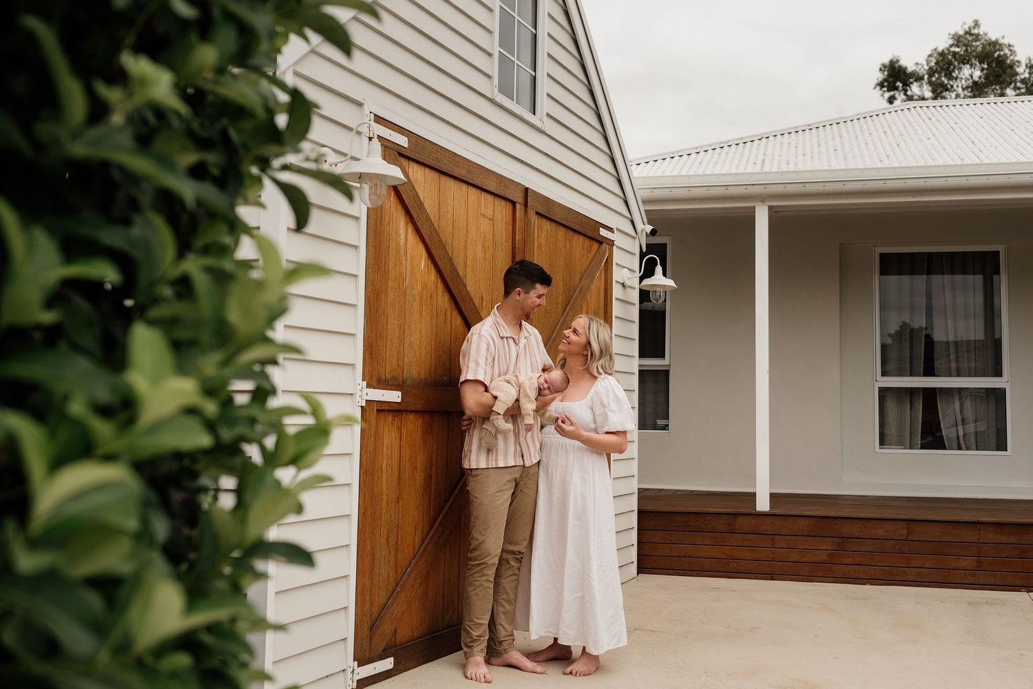 Couple standing barefoot by wooden barn doors at a white farmhouse, holding a bouquet in a romantic outdoor setting.