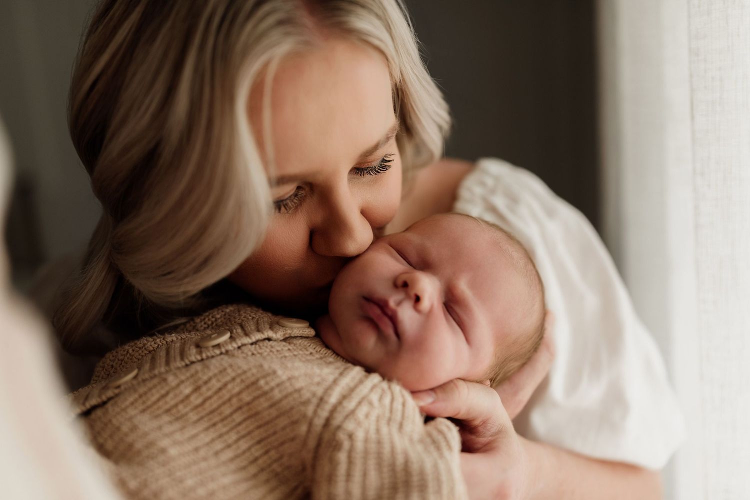 A tender moment as a blonde woman gently kisses a sleeping newborn baby wrapped in a cozy knit blanket.