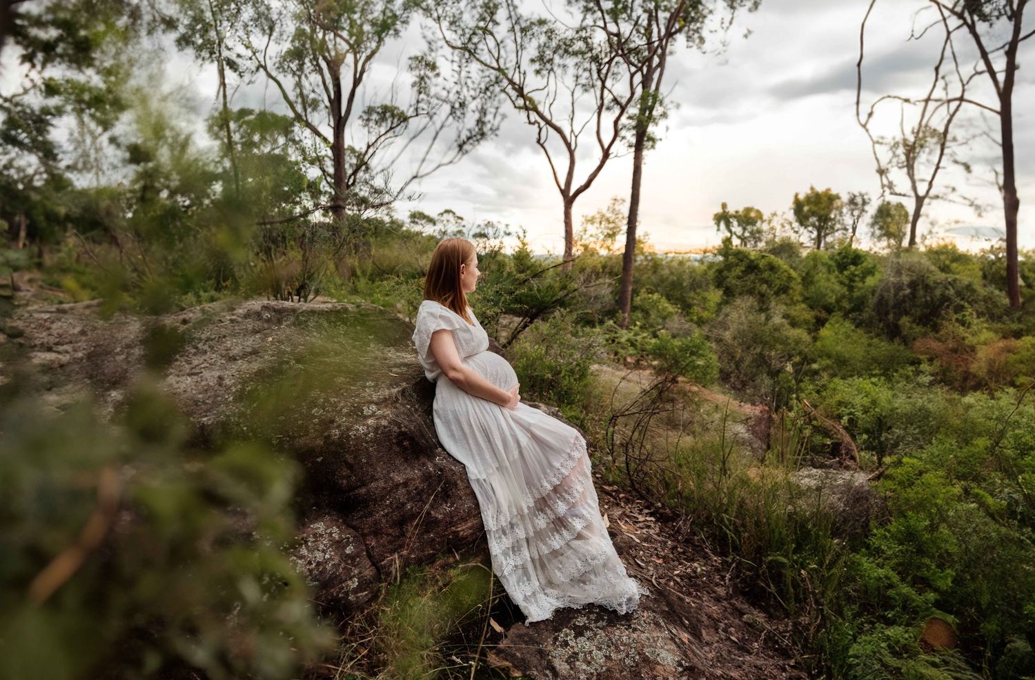 Pregnant woman in white lace dress sits peacefully in Australian bush landscape at dusk.