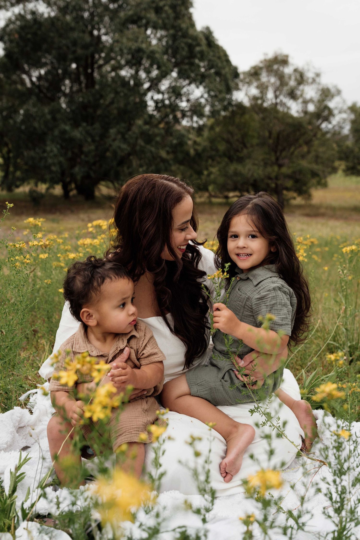 A mother sits on a white blanket in a wildflower field with her two young children, surrounded by yellow blooms and trees.