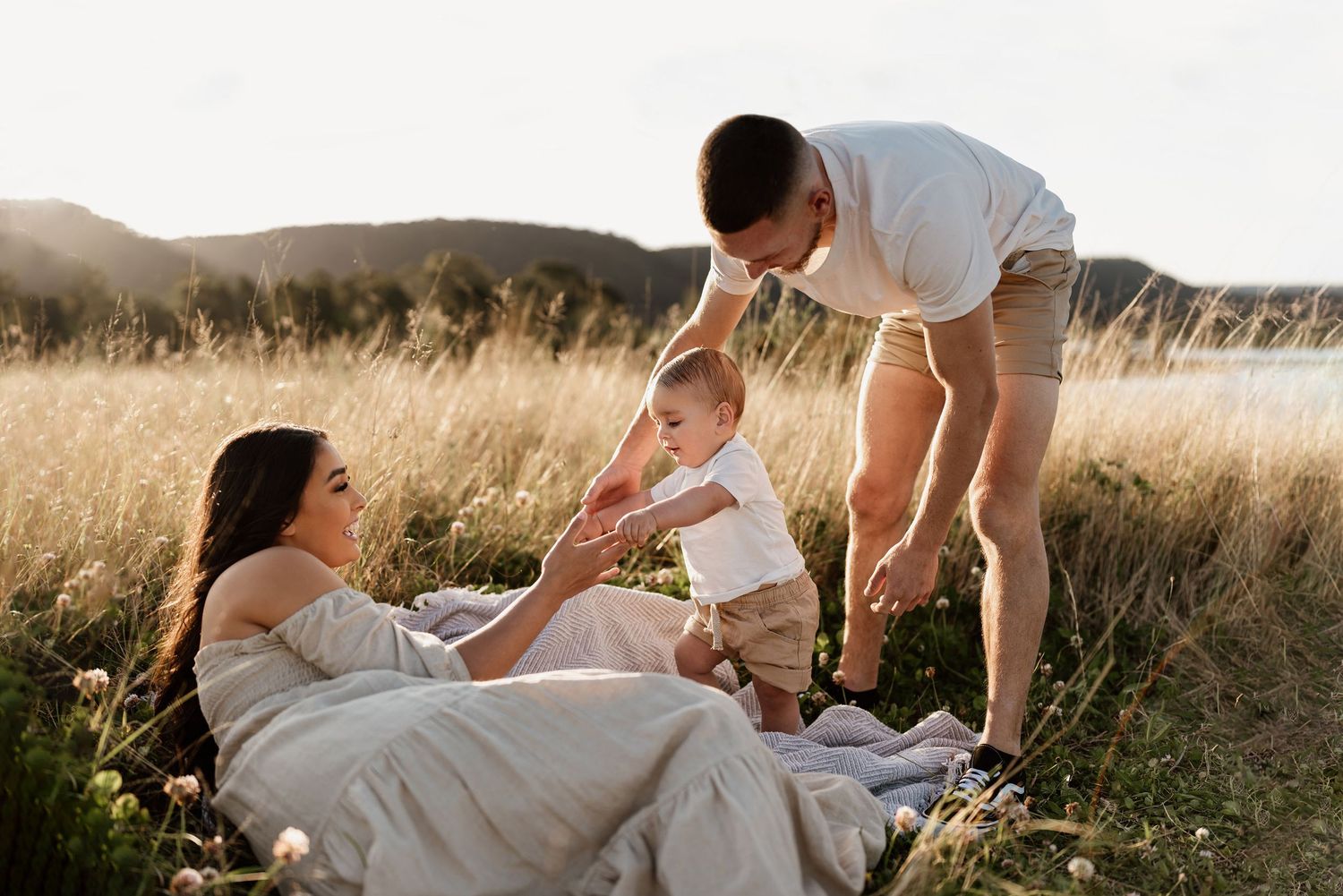 Young family relaxing in golden grass field with father helping baby stand near mother.