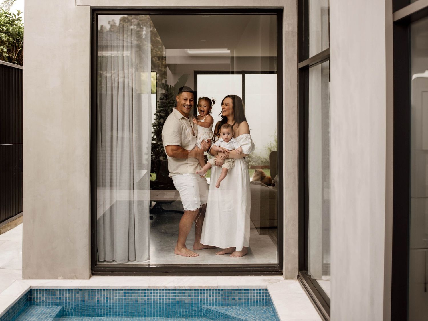 Happy family of four smiling in glass doorway of contemporary home with pool.