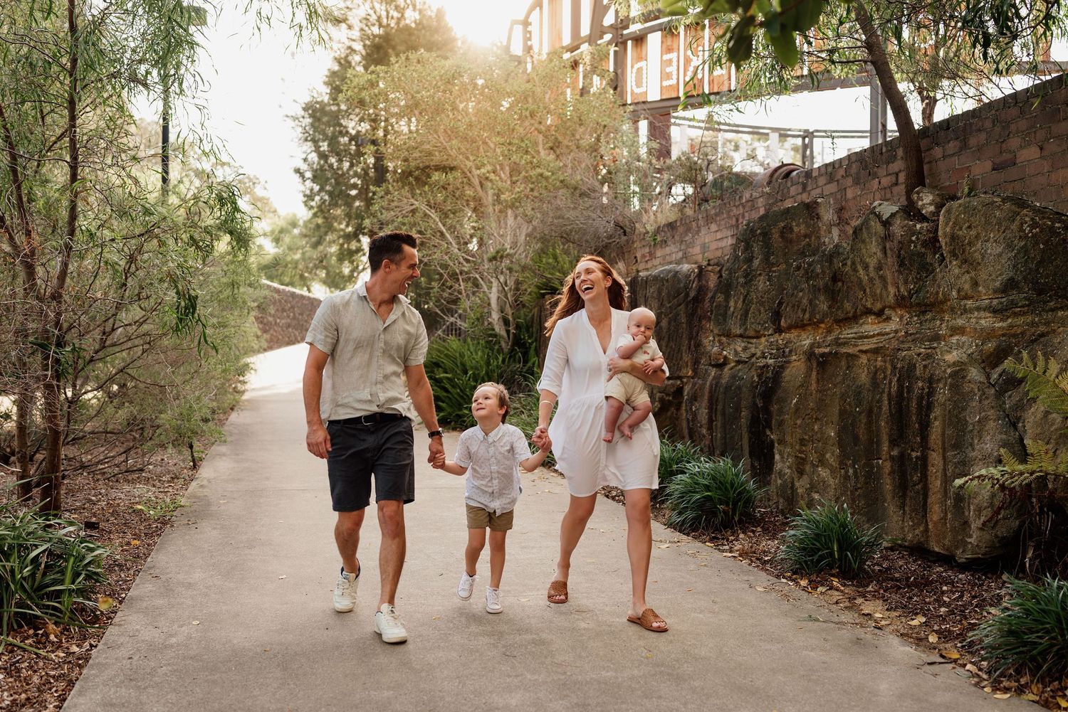 Happy family of four walking along a sunny tree-lined path in casual summer outfits.