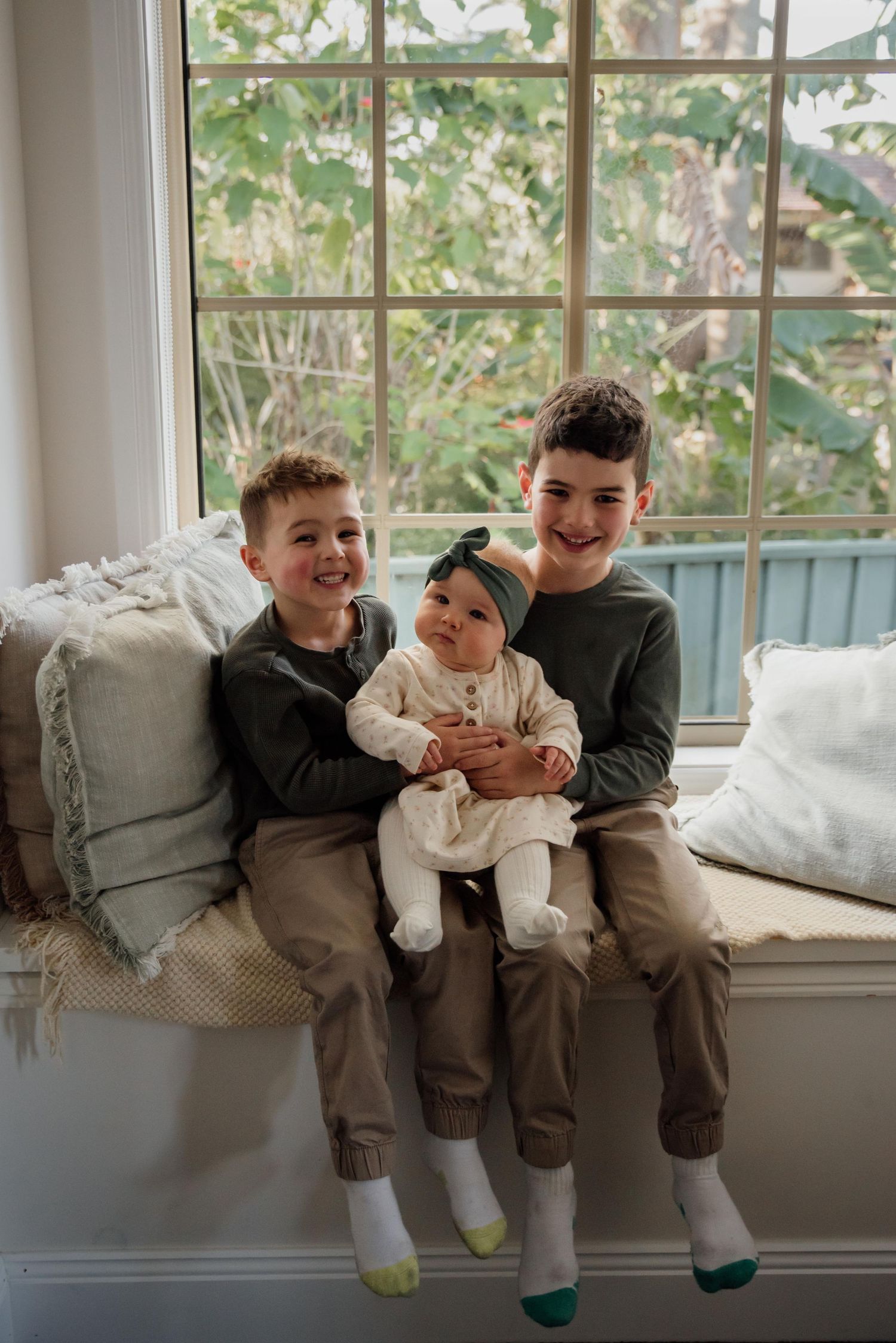 Two smiling boys in matching green shirts and khakis sit on a window seat holding a baby girl in a white outfit.