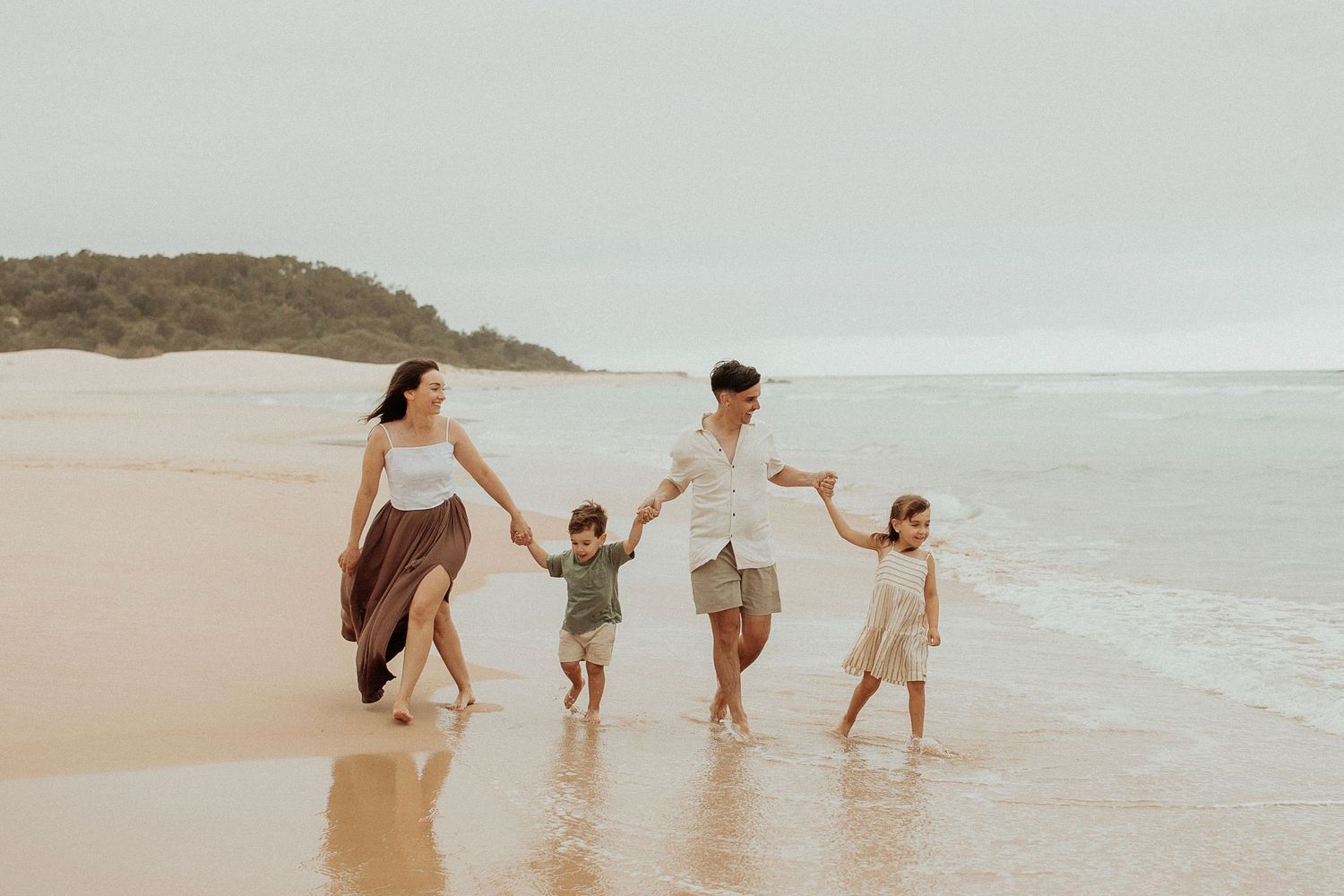Happy family of four holding hands walking along sandy beach shoreline together.