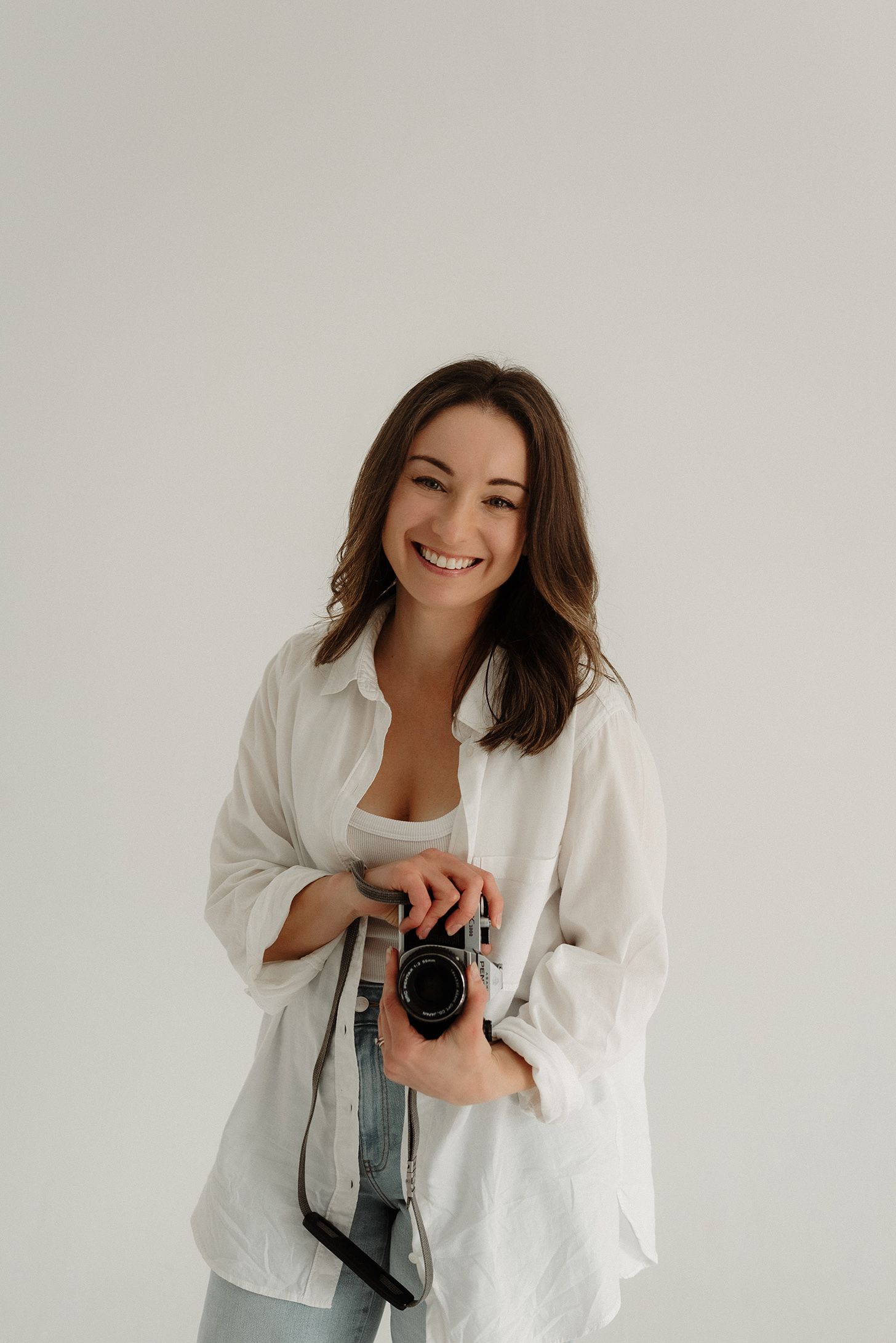 Smiling female photographer in white shirt holding a film camera against a light background.
