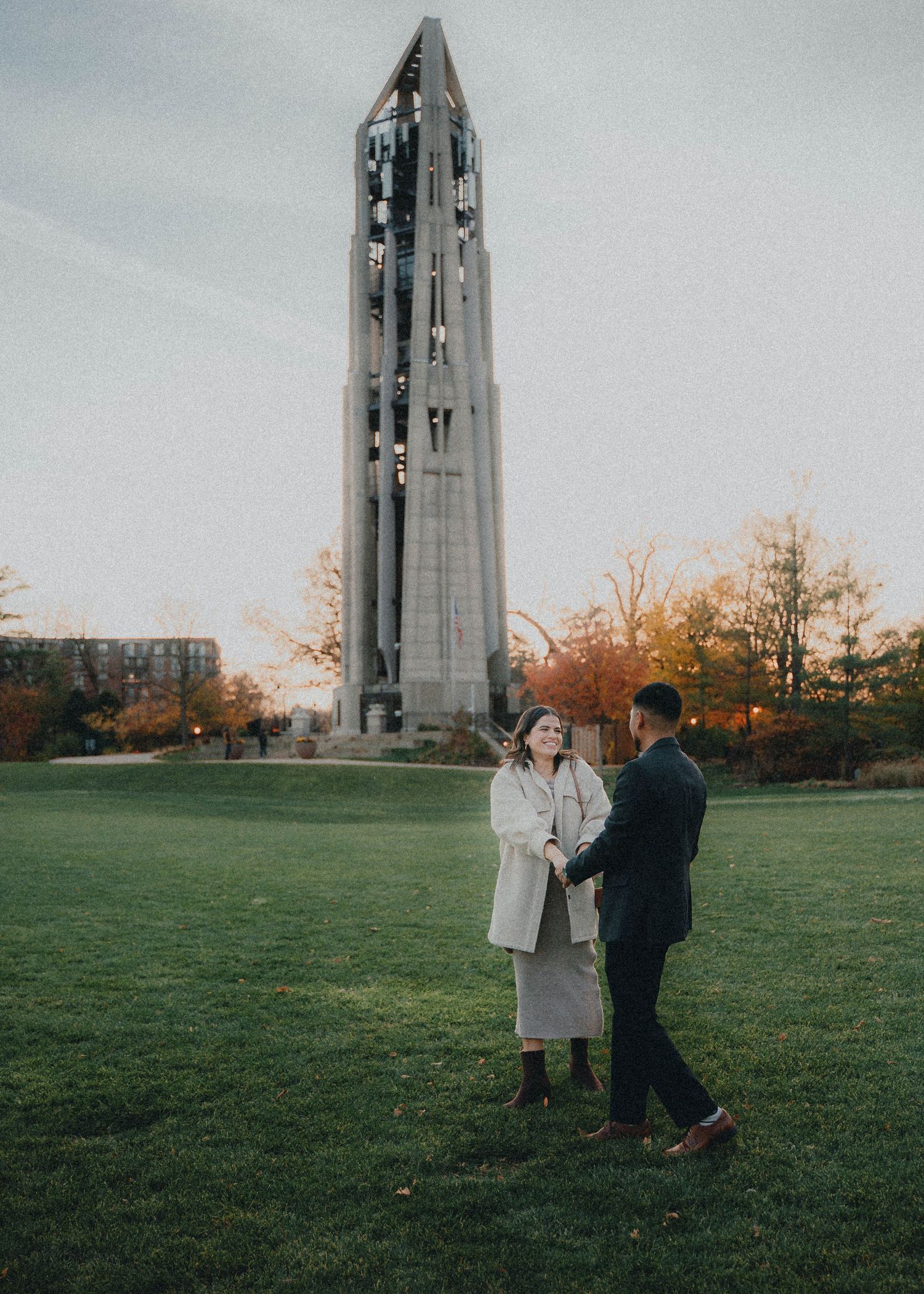 A couple shares a moment on a lawn in front of a tall stone bell tower during autumn.