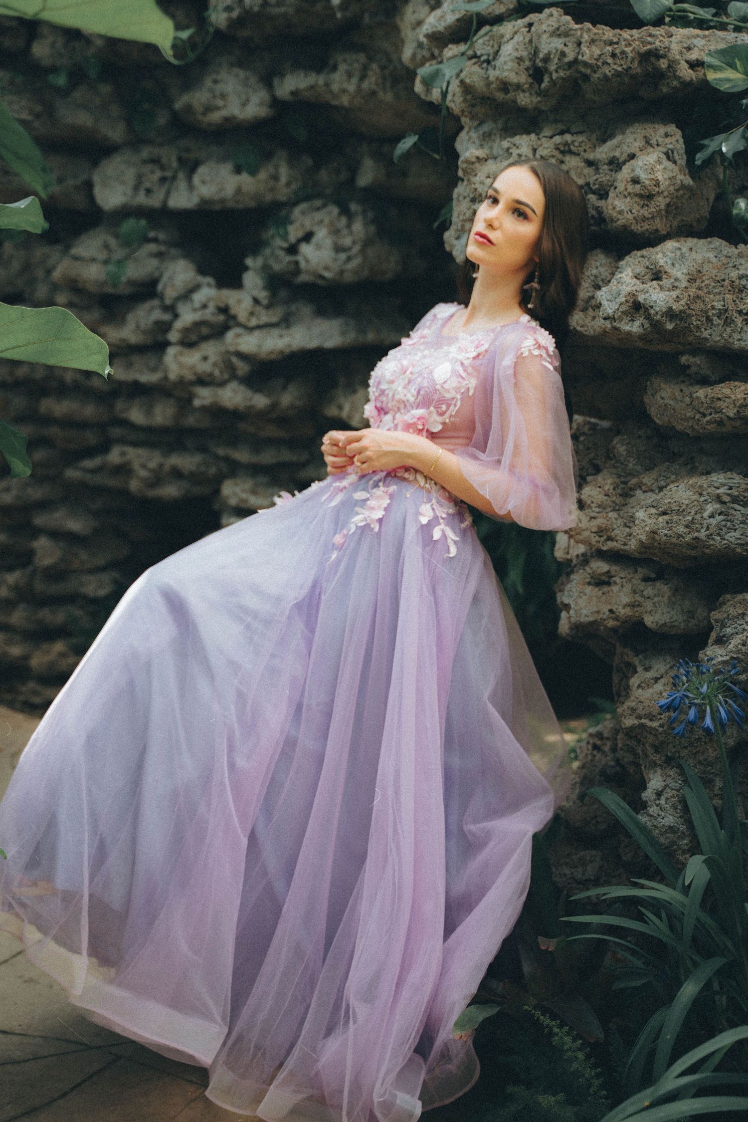 Romantic photoshoot showcasing a flowing lavender gown with pink embellishments against rustic stone walls.