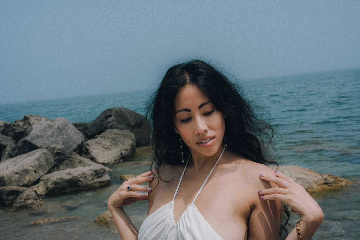 Young woman in white halter dress poses by rocky seaside with windswept dark hair on a misty day.