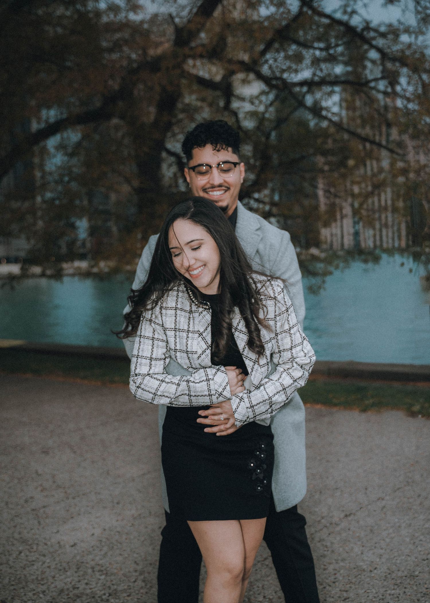 A couple shares a playful moment by a lake on an autumn day, with the person in a plaid shirt embracing their partner.